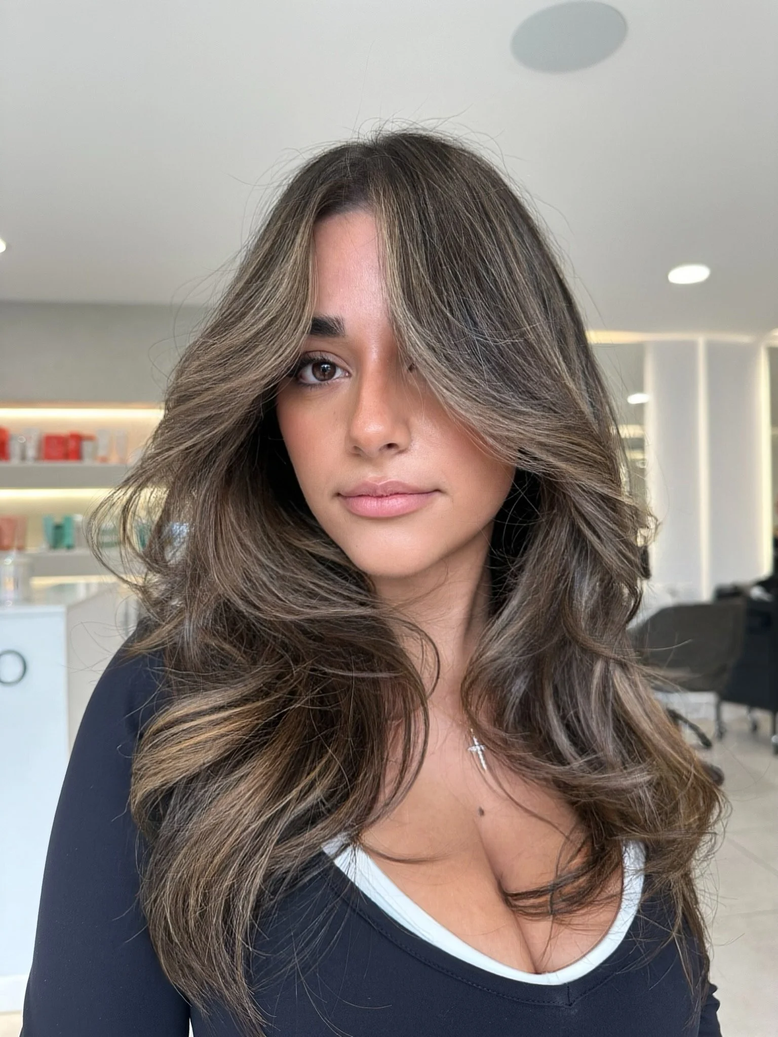 Portrait of a woman with long wavy brown hair and side-swept bangs, wearing a black top with a white lining and a silver cross necklace, in a modern interior space with shelves and seating.