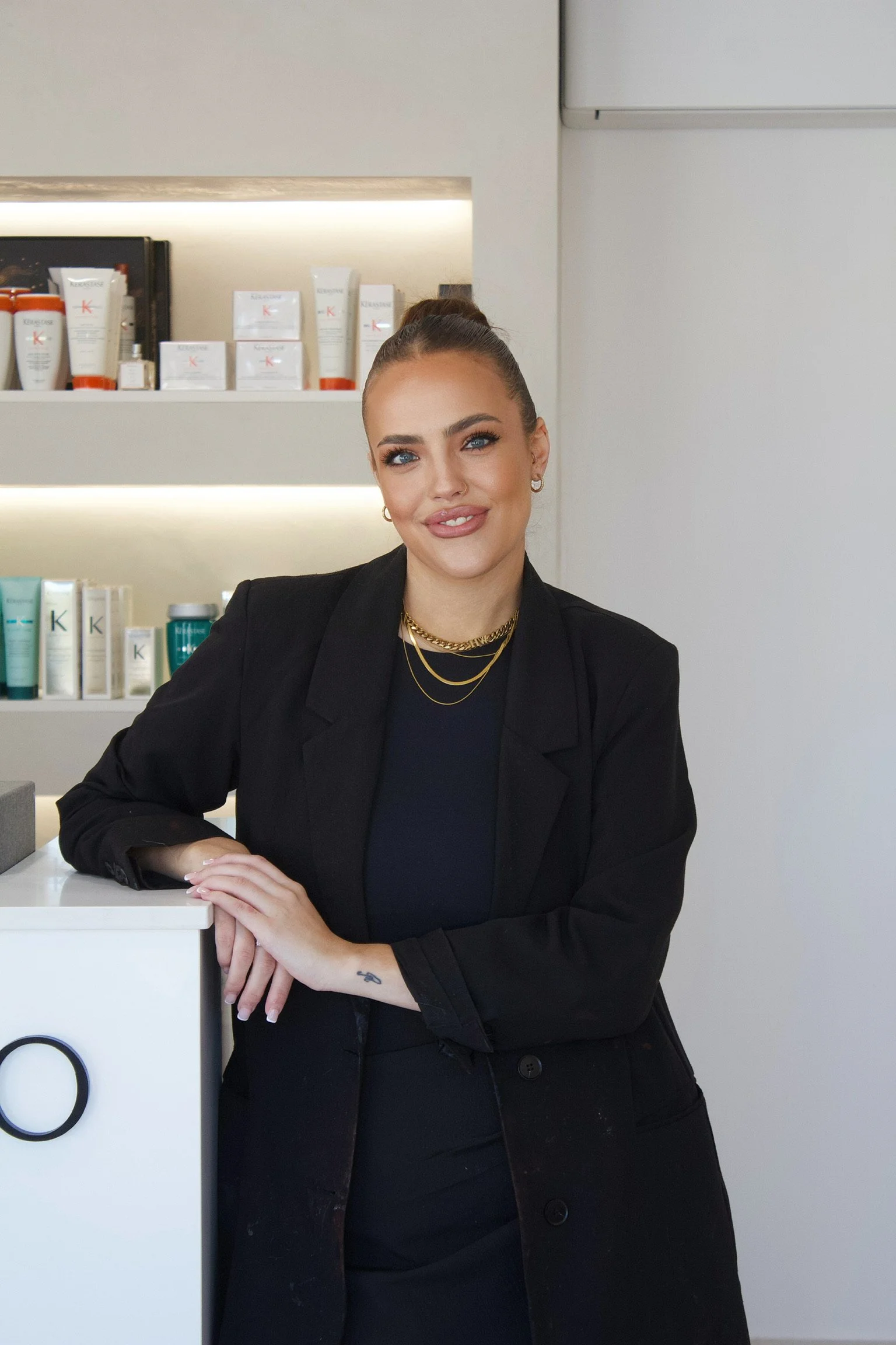 A woman with styled hair and makeup, wearing a black blazer and jewelry, standing inside a skincare or beauty store.