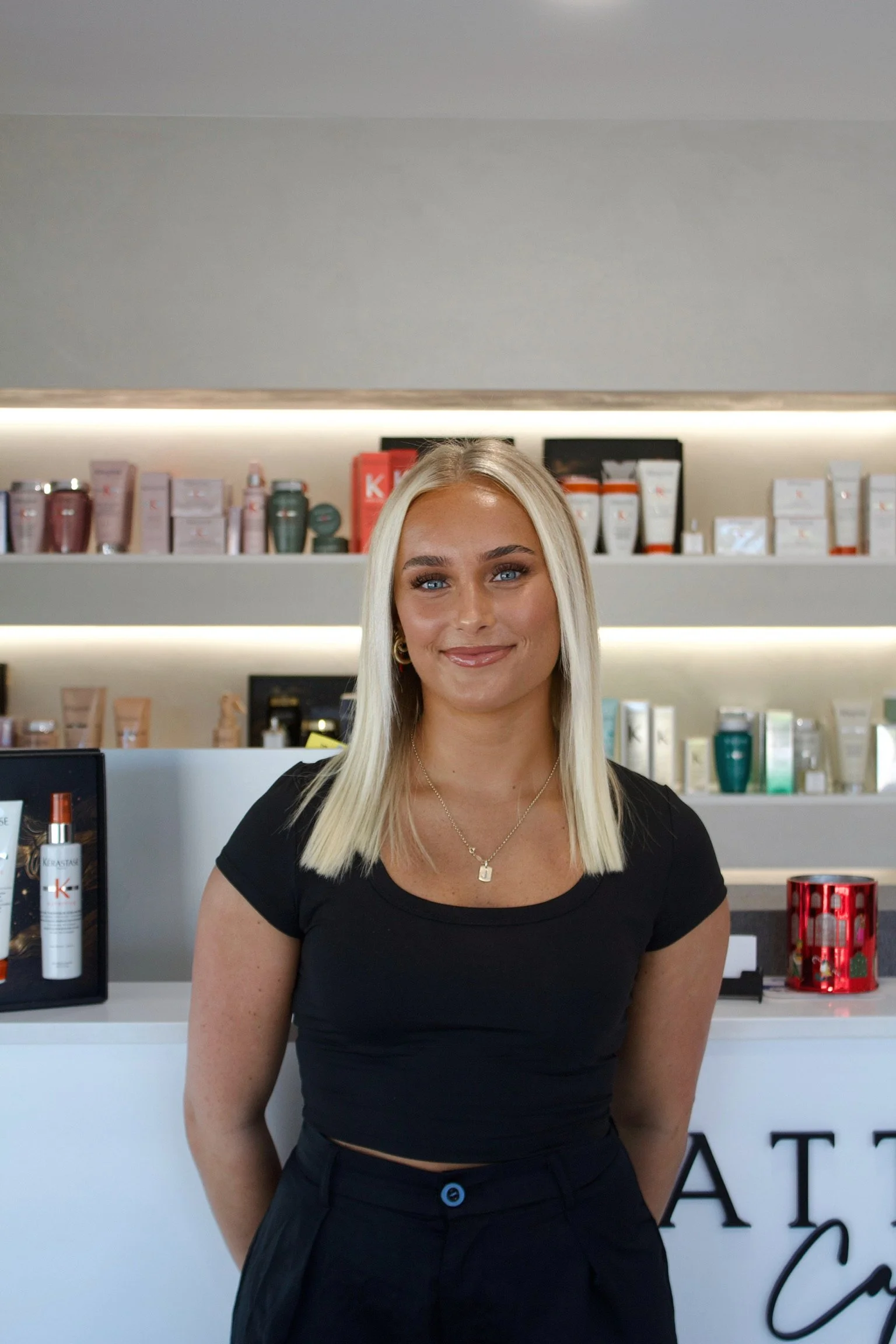 A smiling blonde woman in a black top standing behind a white counter at a retail store with shelves of skincare products in the background.