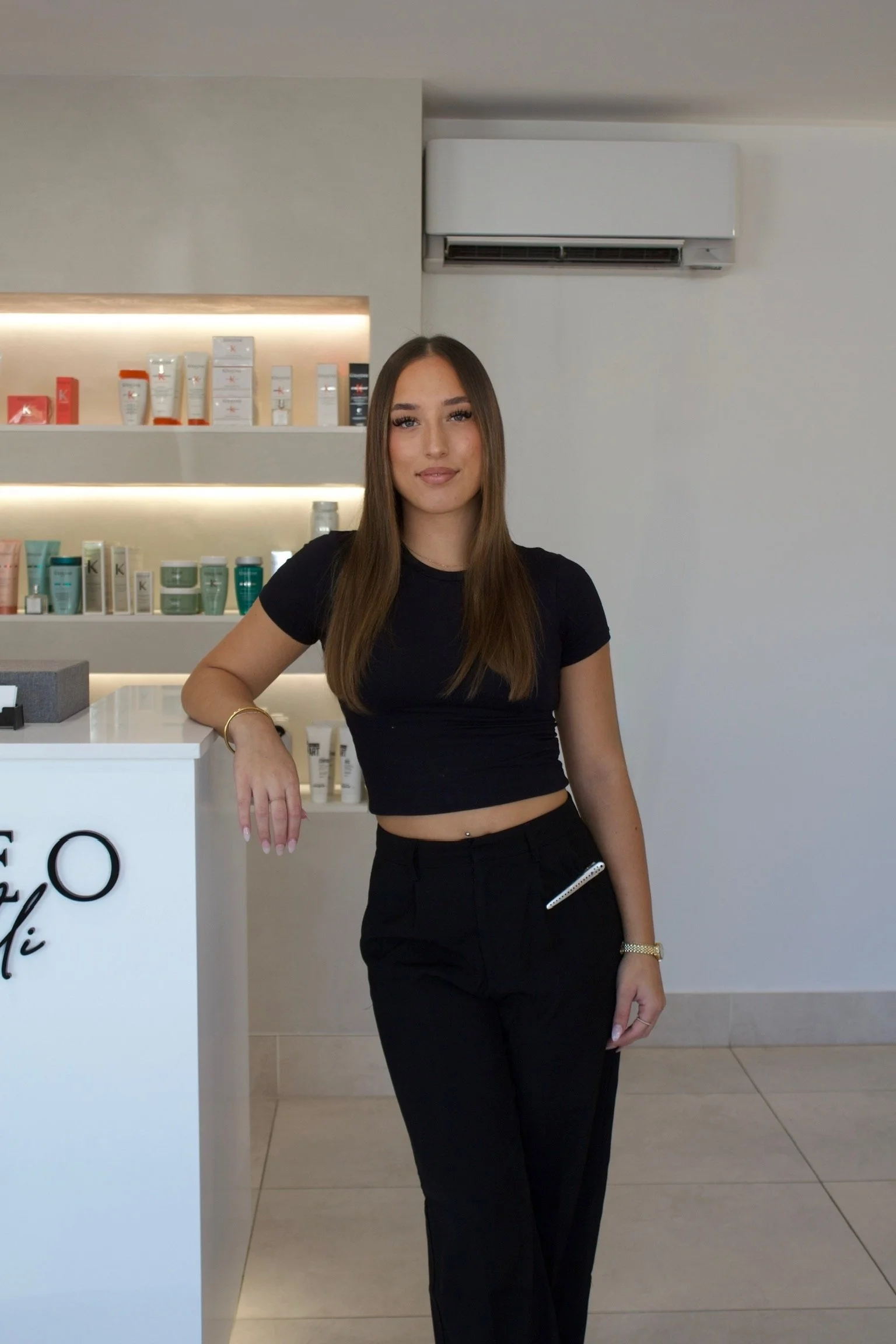Young woman standing indoors next to a white counter with skincare products on shelves behind her.