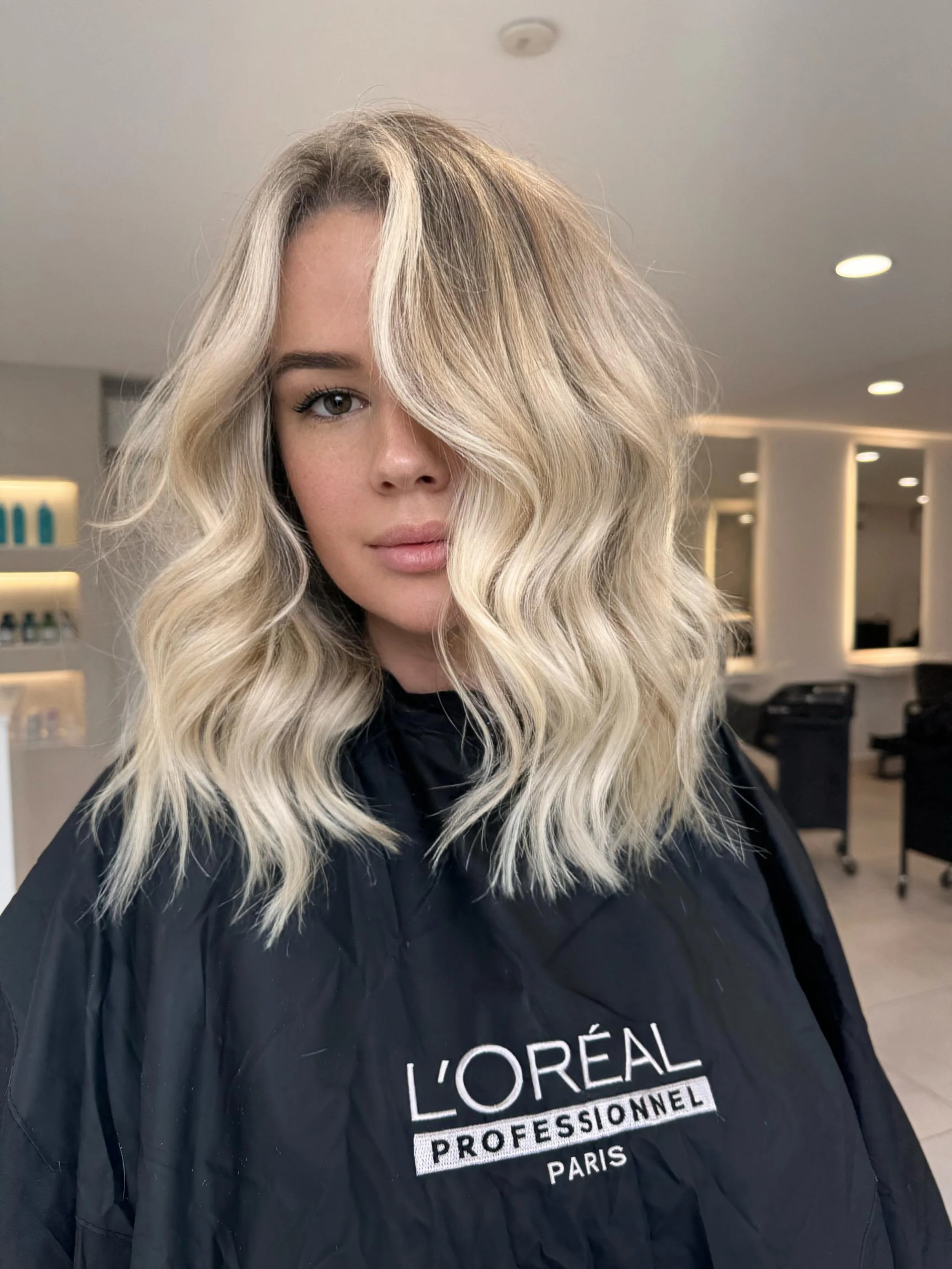 A woman with blonde, shoulder-length wavy hair sitting in a salon, wearing a black cape with 'L'Oréal Professionnel Paris' written on it.