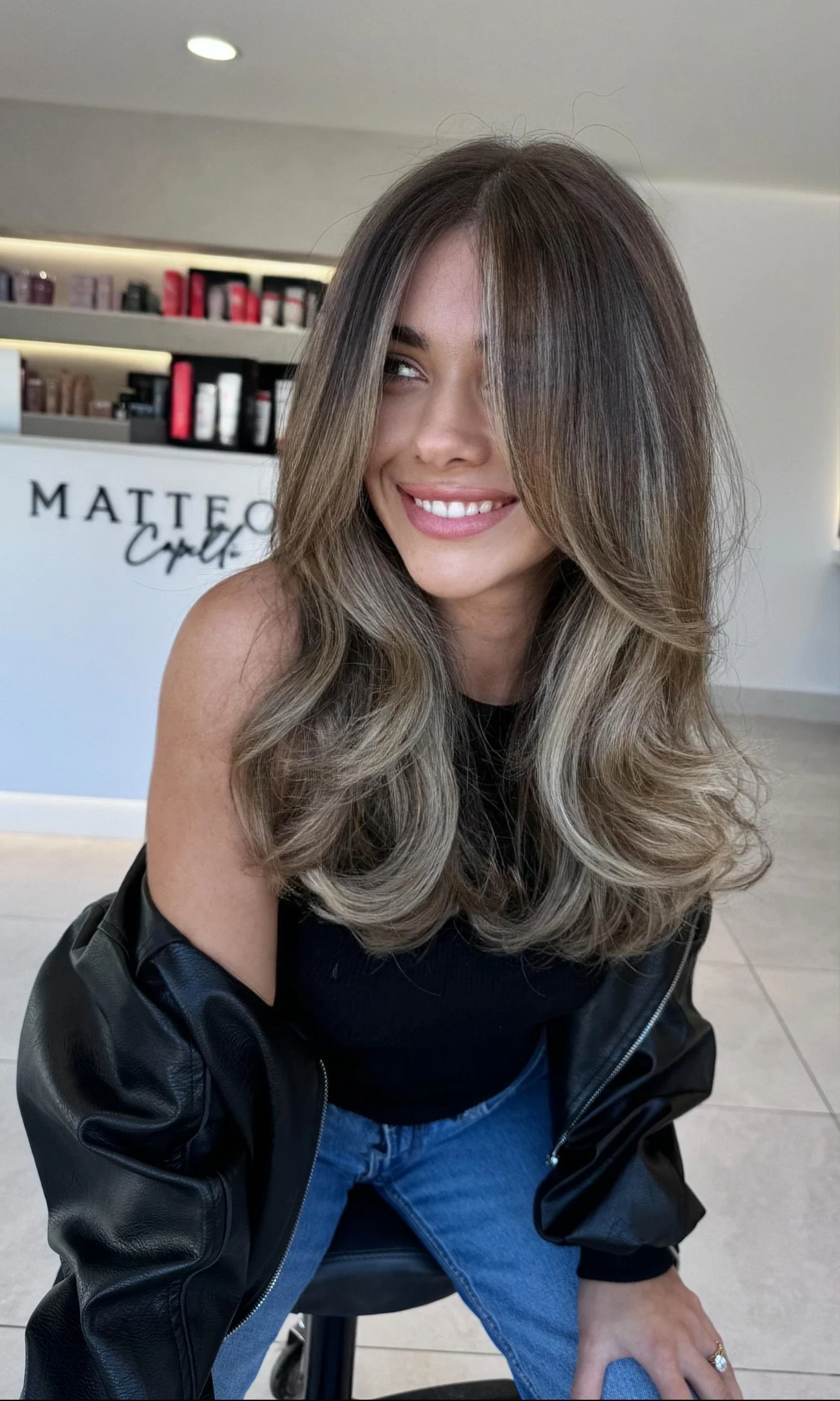 A woman with long, layered hair and a black leather jacket sitting in a salon, smiling and looking to the side.