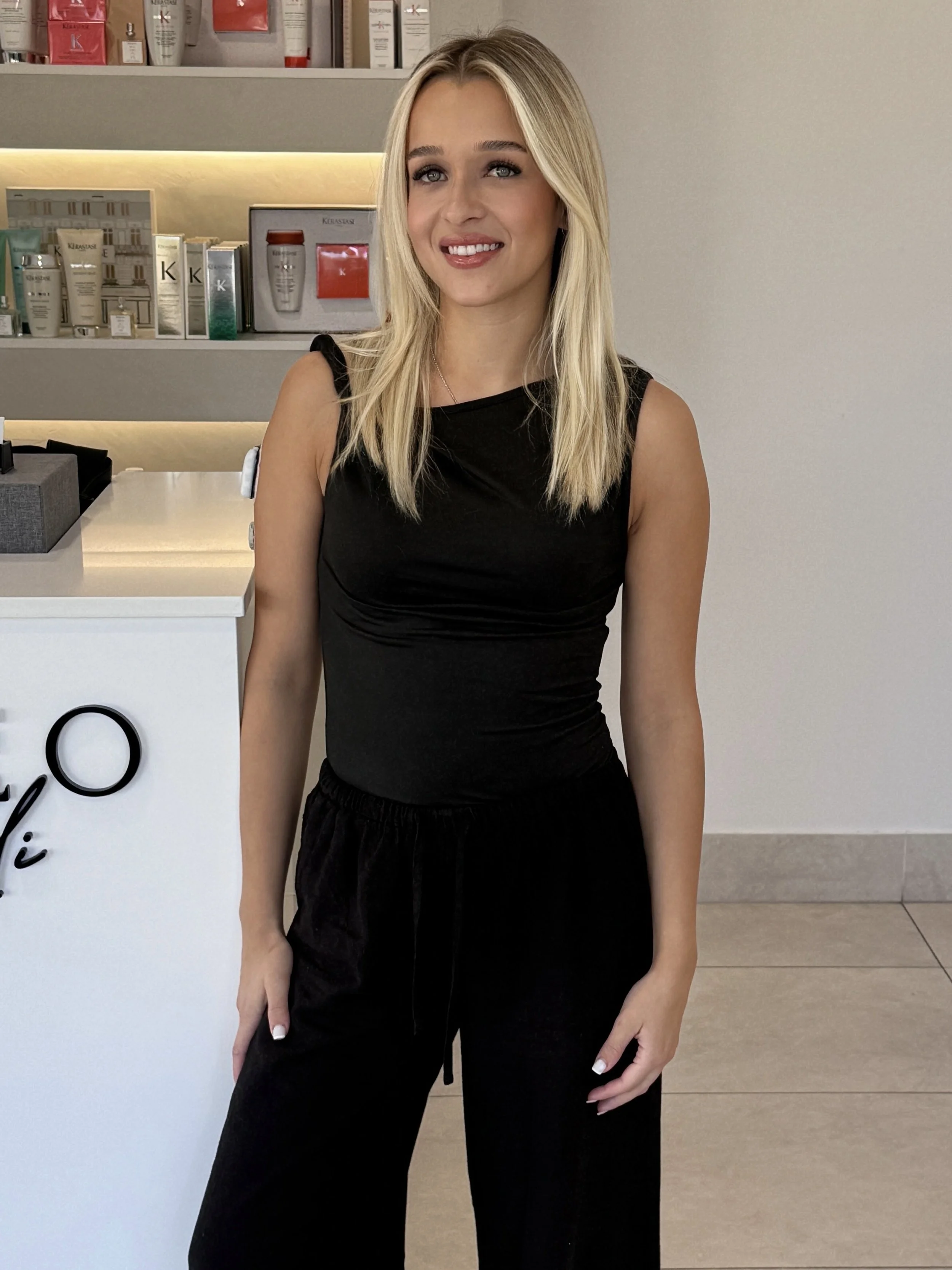 Young woman with long dark hair, wearing a black top and black pants, standing inside a modern store or salon with shelves of skincare or cosmetic products in the background.