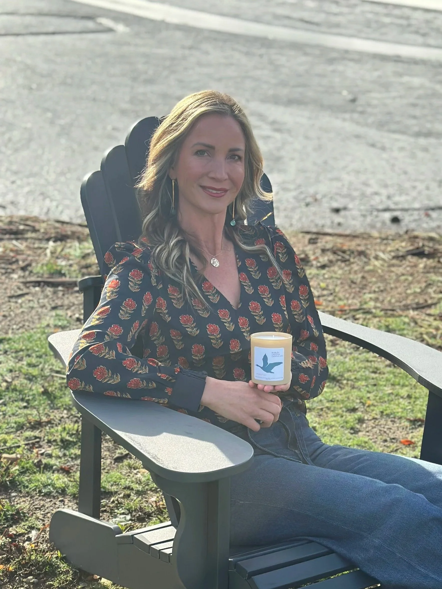 A woman with blonde hair, wearing a floral blouse and blue jeans, sitting in a reclining outdoor chair on grass, holding a white candle with a blue hummingbird design.