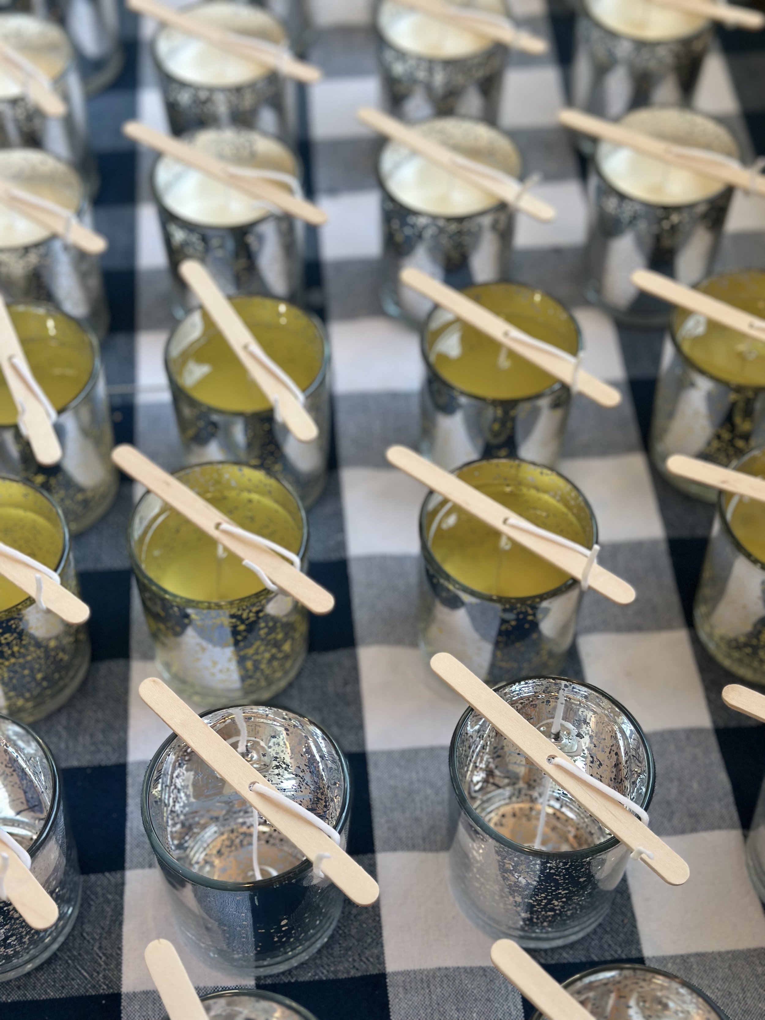 Rows of small glass containers filled with yellow and clear candles, each with a wooden stick for a wick, placed on a black and white checkered tablecloth.