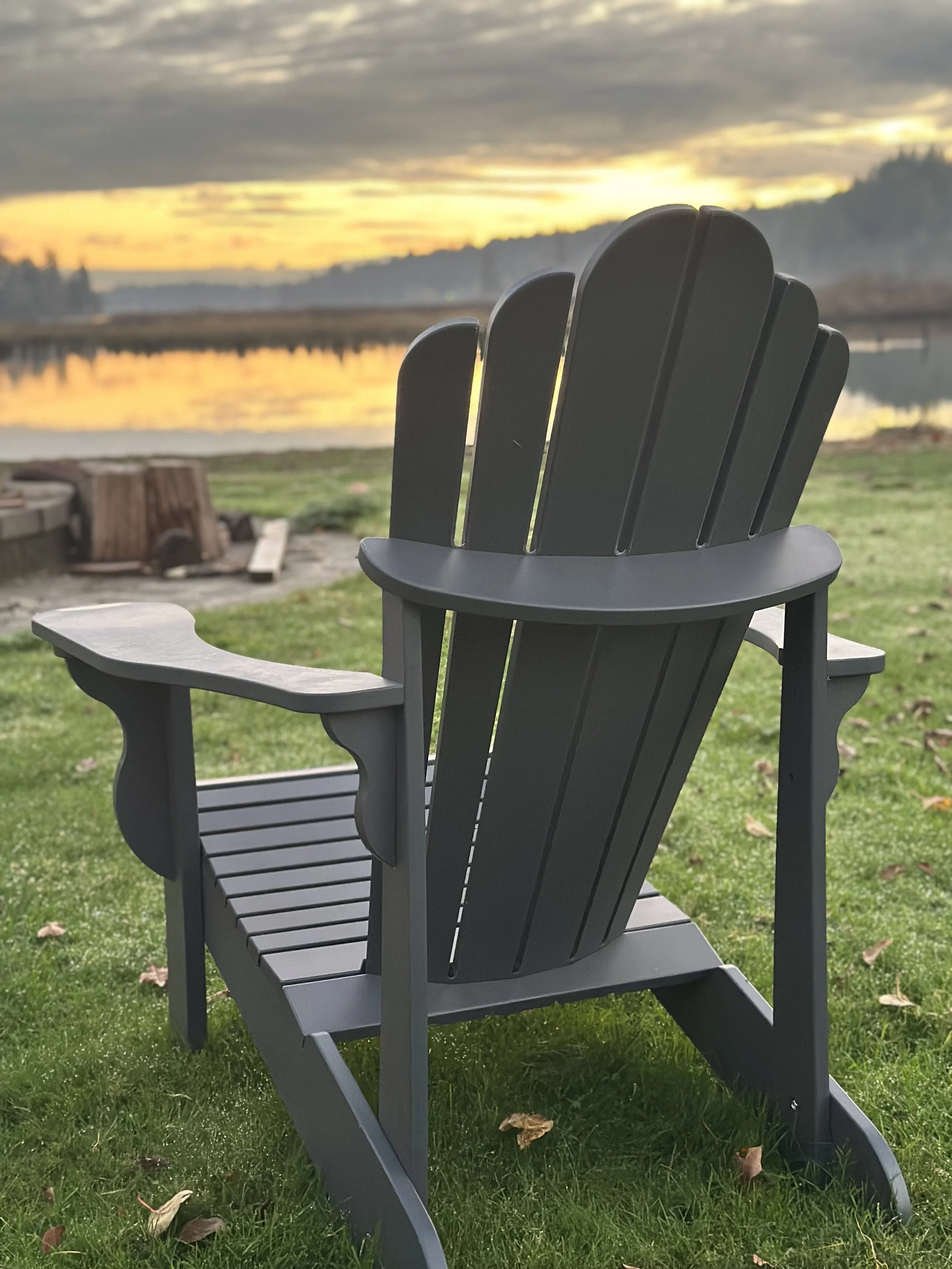 Gray Adirondack chair facing a lake at sunset with a treeline in the distance.