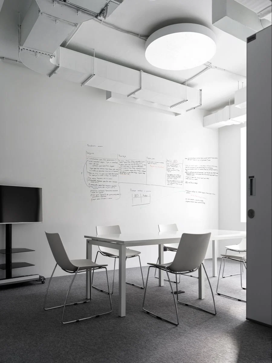 Salle de réunion minimaliste avec une table blanc, quatre chaises blanches, un mur blanc avec écriture manuscrite, un grand plafonnier rond, un téléviseur noir sur un support mobile, et une lumière naturelle provenant de la fenêtre.