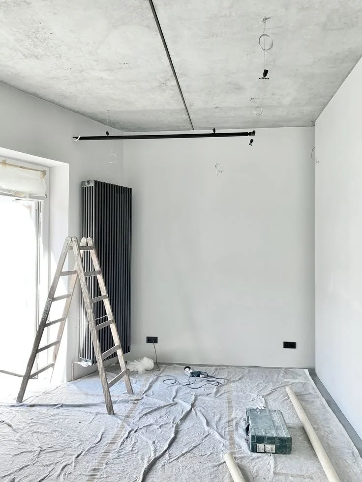 Une pièce en cours de rénovation avec un plafond en béton apparent, un radiateur noir vertical, une échelle en bois, des câbles électriques, un sac blanc et des tuyaux en plastique sur le sol.