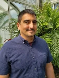 Man with short dark hair, mustache, and goatee, wearing a blue button-up shirt, standing in front of green plants in a greenhouse.