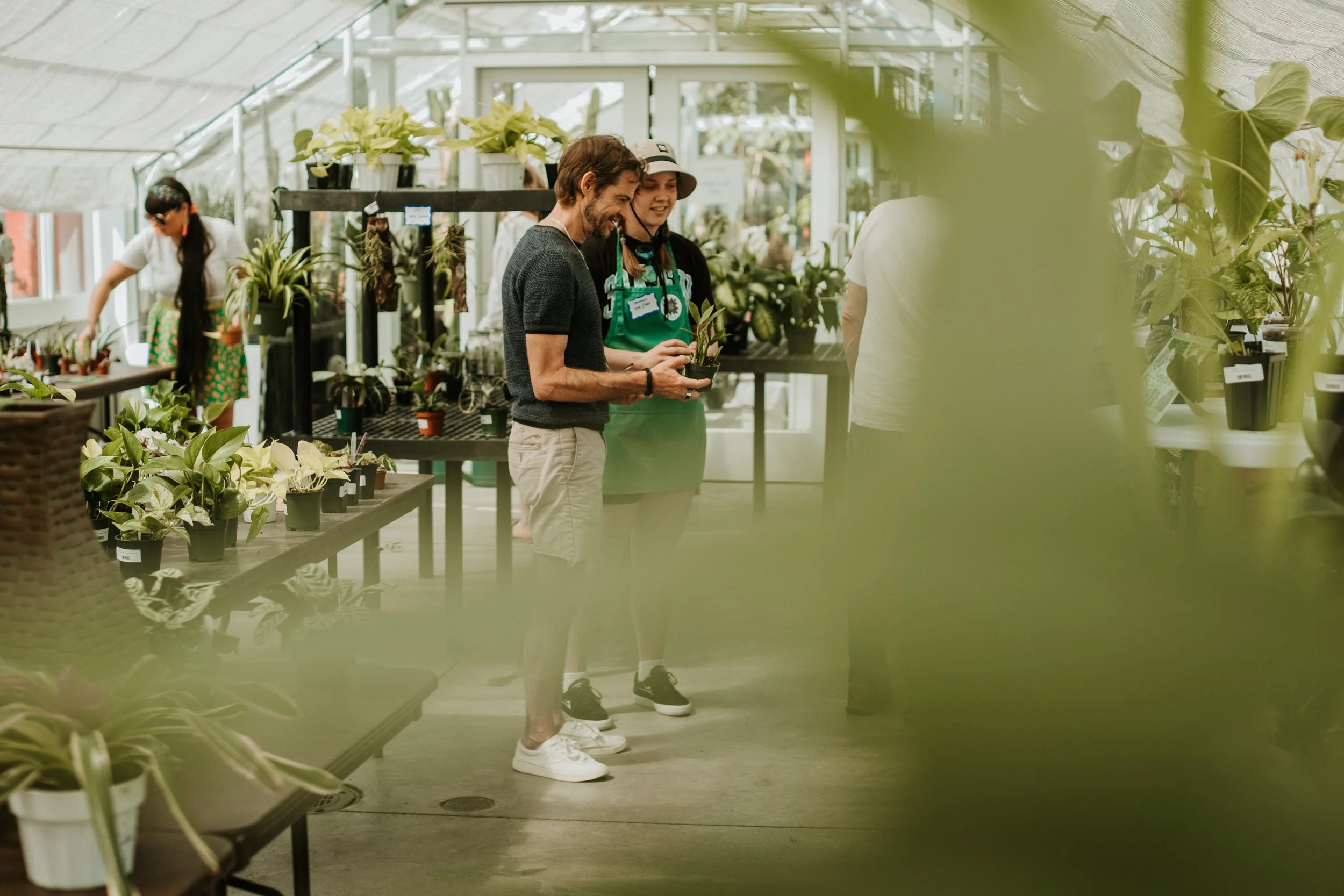 People shopping for plants inside a greenhouse or garden center, with a man and woman examining a potted plant, surrounded by various potted plants on shelves and tables.