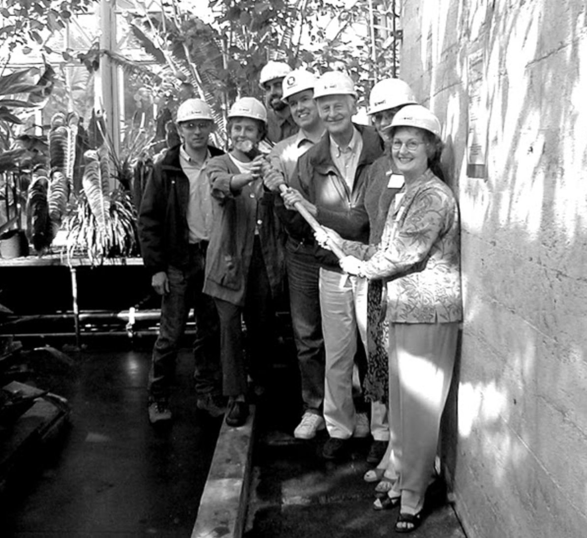A group of seven people wearing construction helmets, gathered inside a greenhouse or botanical space, holding a large pair of scissors for a ribbon-cutting ceremony.