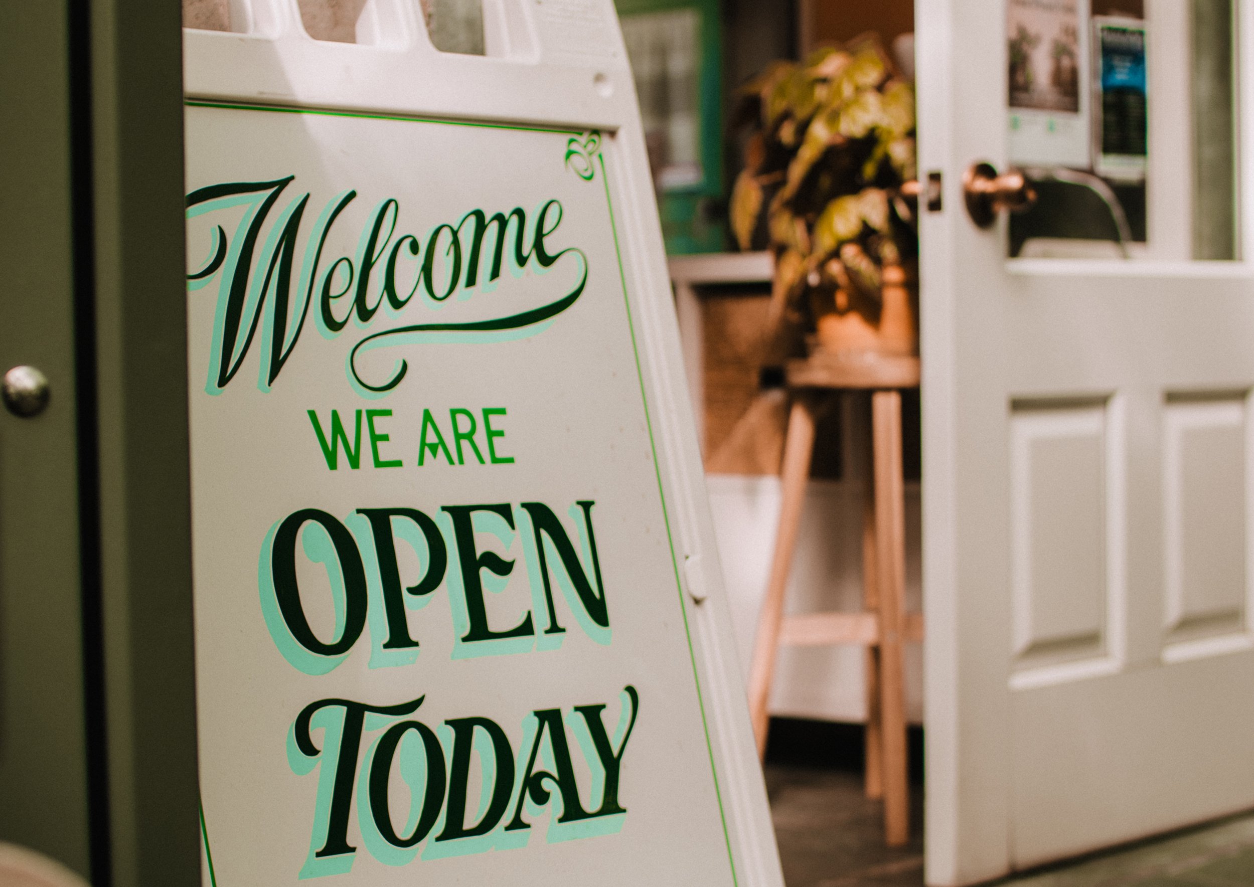 A white signboard with green and black lettering that says "Welcome, We Are Open Today," placed outside a shop or cafe entrance. Part of the door and interior with plants and decorations are visible in the background.