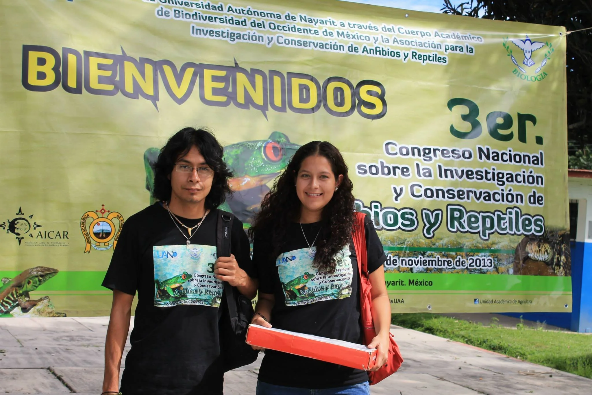 Dos jóvenes con camisetas negras con logotipo y texto del congreso posando frente a un cartel grande en un evento académico sobre investigación y conservación de anfibios y reptiles. La mujer sostiene una caja y ambos llevan mochila. El cartel tiene texto en español y logotipos de instituciones.