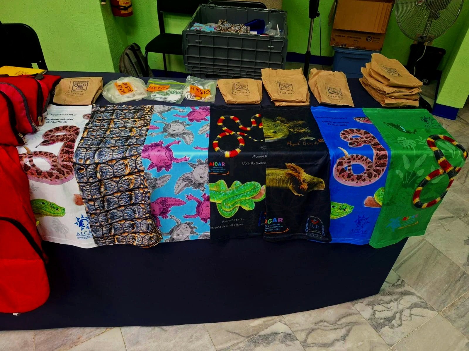 Mesa con exhibiciones de bandanas con temas de reptiles, incluyendo serpientes y lagartos, de diferentes colores y patrones, y bolsas de papel y plástico con etiquetas de precios.
