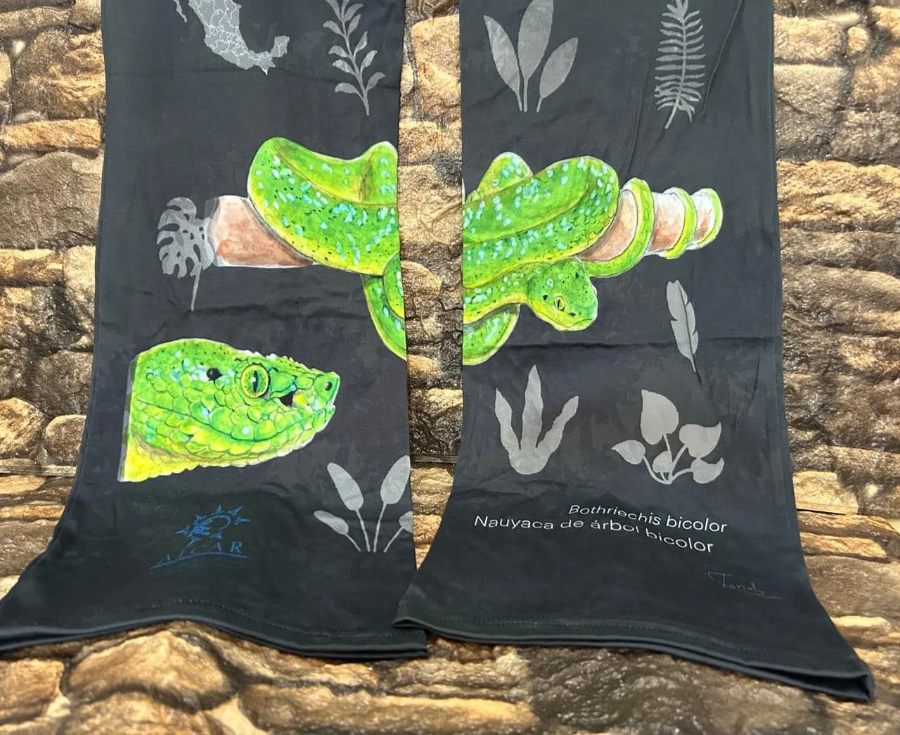 Tela negra con ilustraciones de una serpiente de árbol bicolor y una cabeza de reptil, acompañada por hojas en color gris. Texto en la tela indica «Bothriechis bicolor» y «Nauyaca de árbol bicolor».