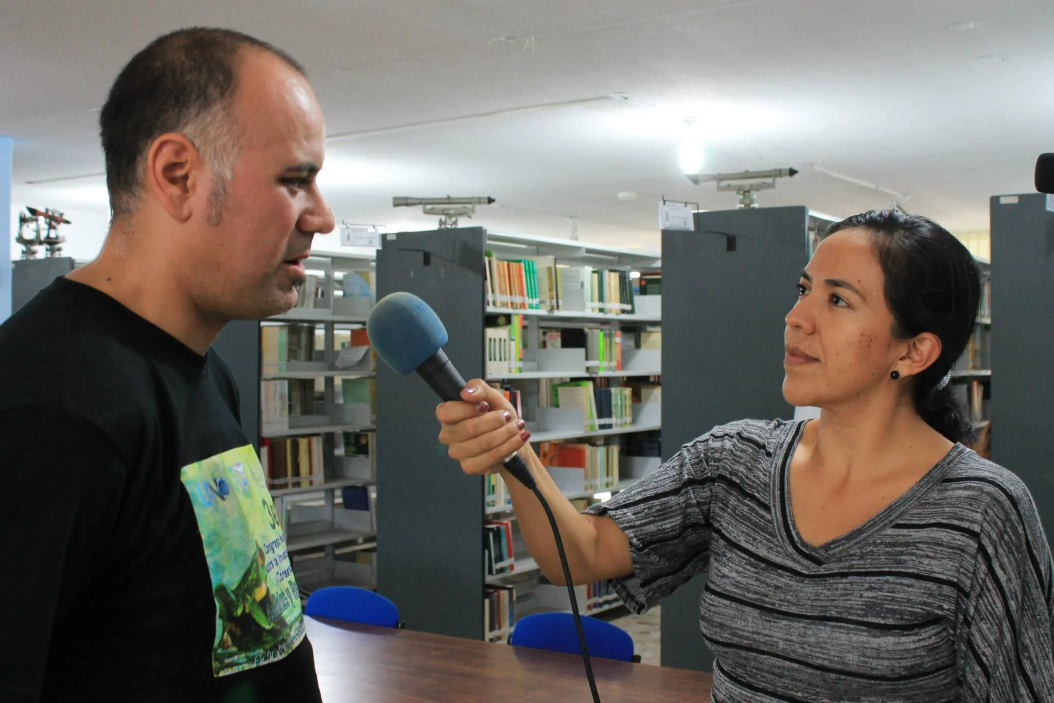 Una mujer realiza una entrevista a un hombre que lleva puesta una camiseta negra, en un entorno de biblioteca con estanterías llenas de libros.