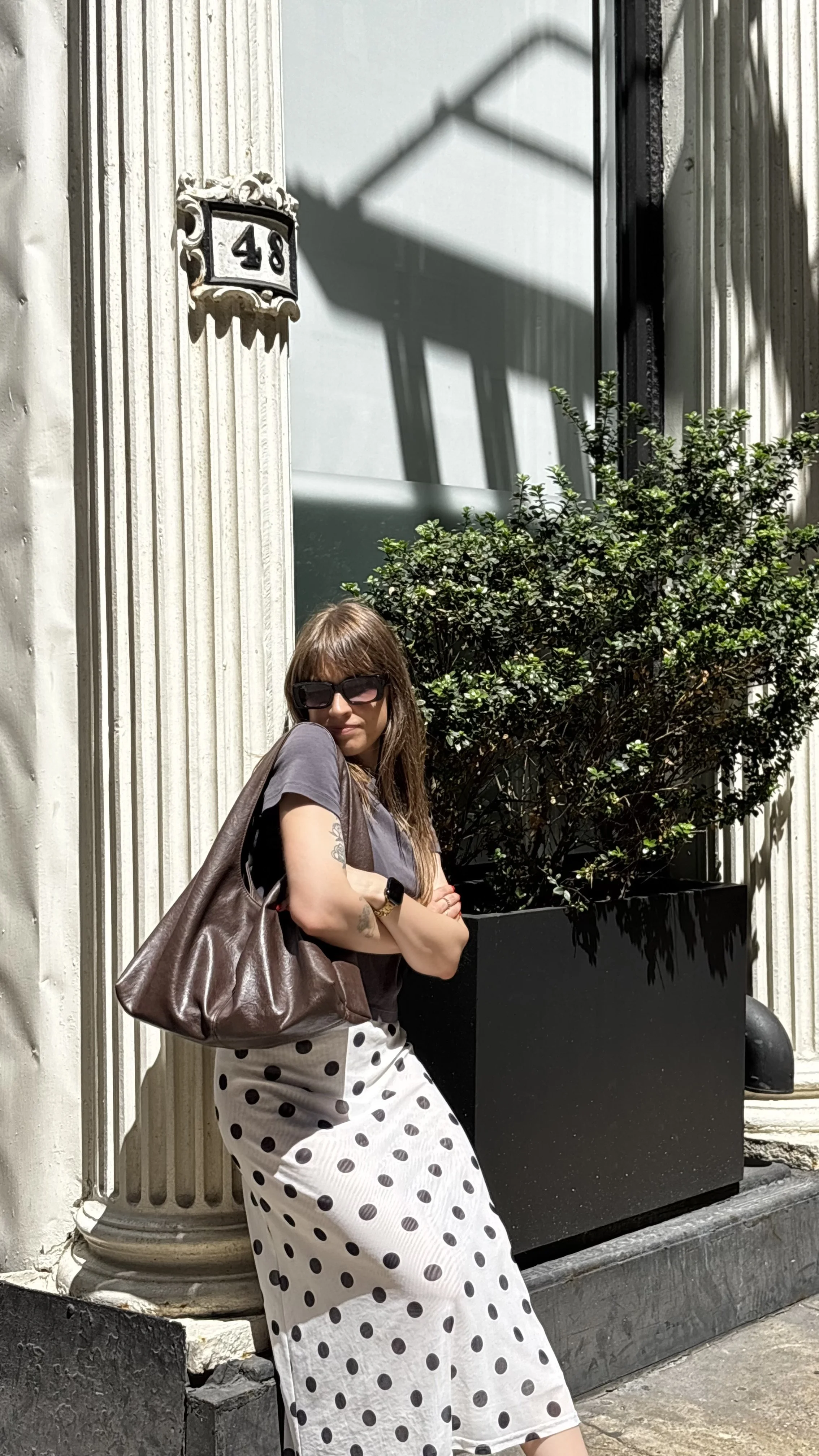 Woman with sunglasses and tattoos leaning against a building with a column, wearing a polka-dot skirt and holding a large brown bag, standing near a potted plant with a shadow of a streetlamp on the wall.