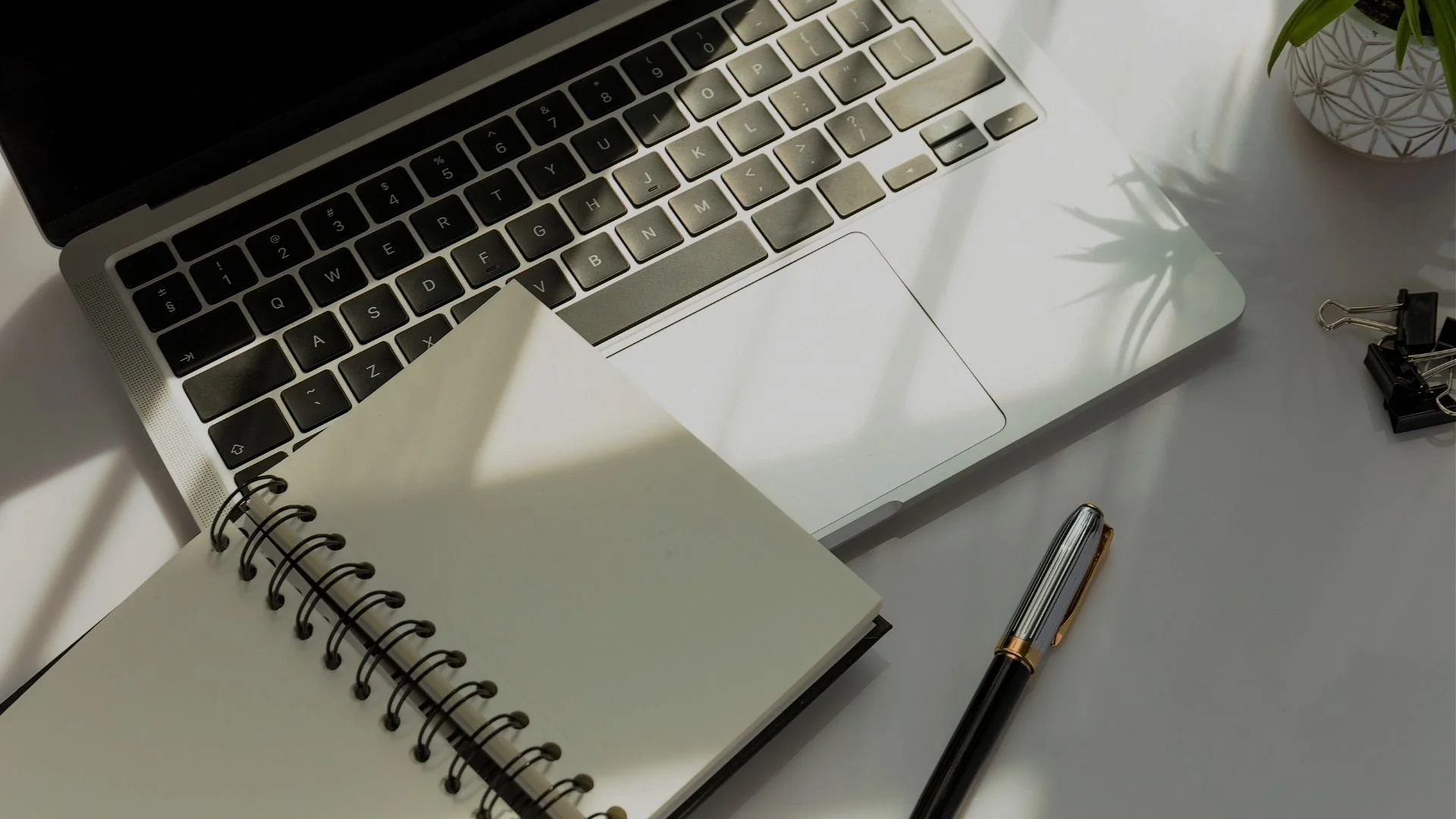 Close-up of a laptop keyboard with a notepad, pen, and binder clips on a desk with a plant in the background.