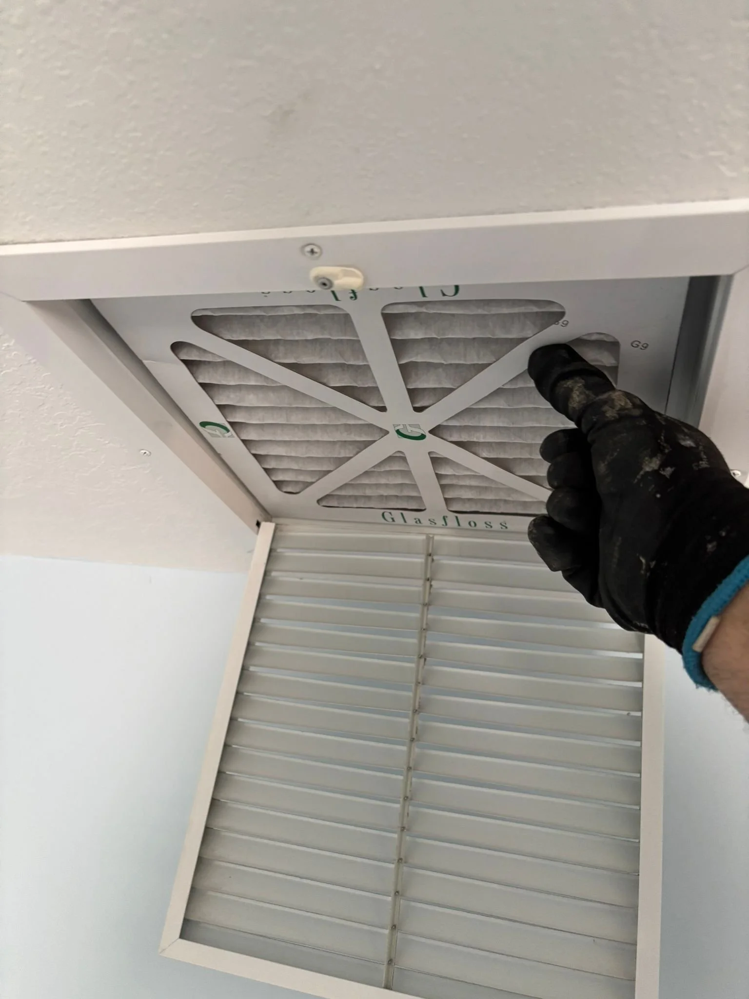 Person wearing a black work glove opening a ceiling vent with a filter inside.