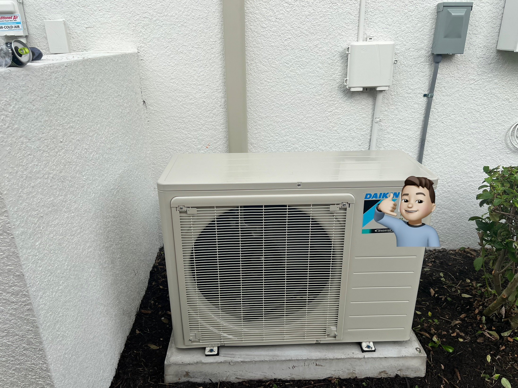 An outdoor air conditioning unit placed on a concrete pad next to a white textured wall, with utility boxes and pipes on the wall behind it. A cartoon character sticker is on the unit, making a thumbs-up gesture.