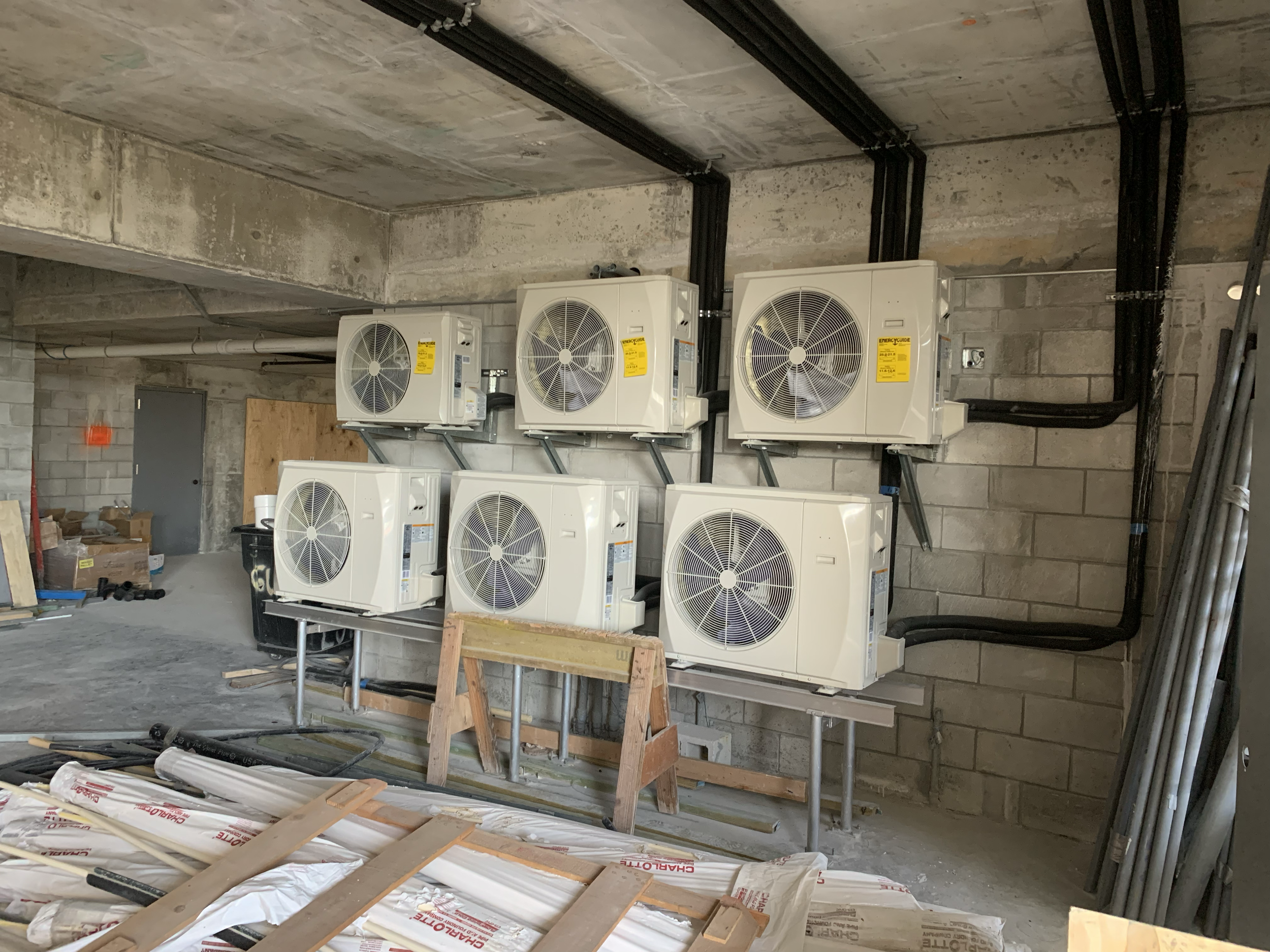 Construction site with six large HVAC units mounted on wall, work materials, and unfinished concrete and brick walls.