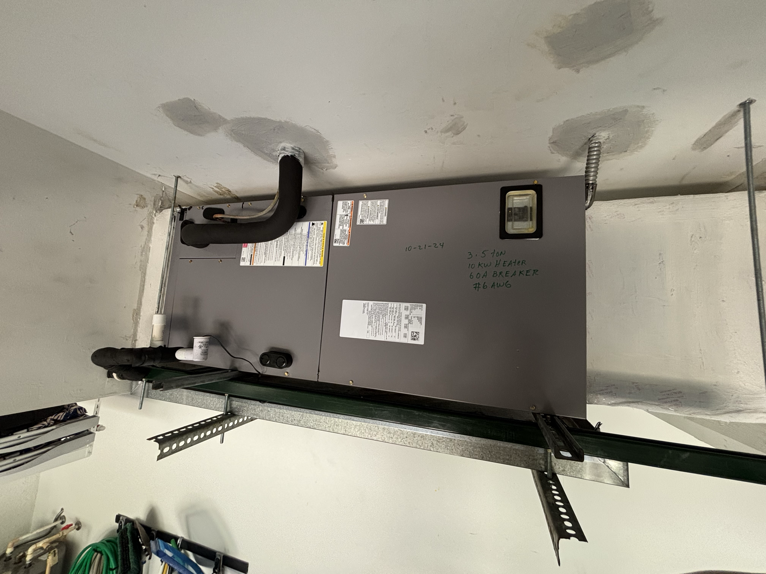 An HVAC unit mounted on a wall in a utility or mechanical room, with pipes and wiring connected to it, and handwritten notes on the front of the unit.