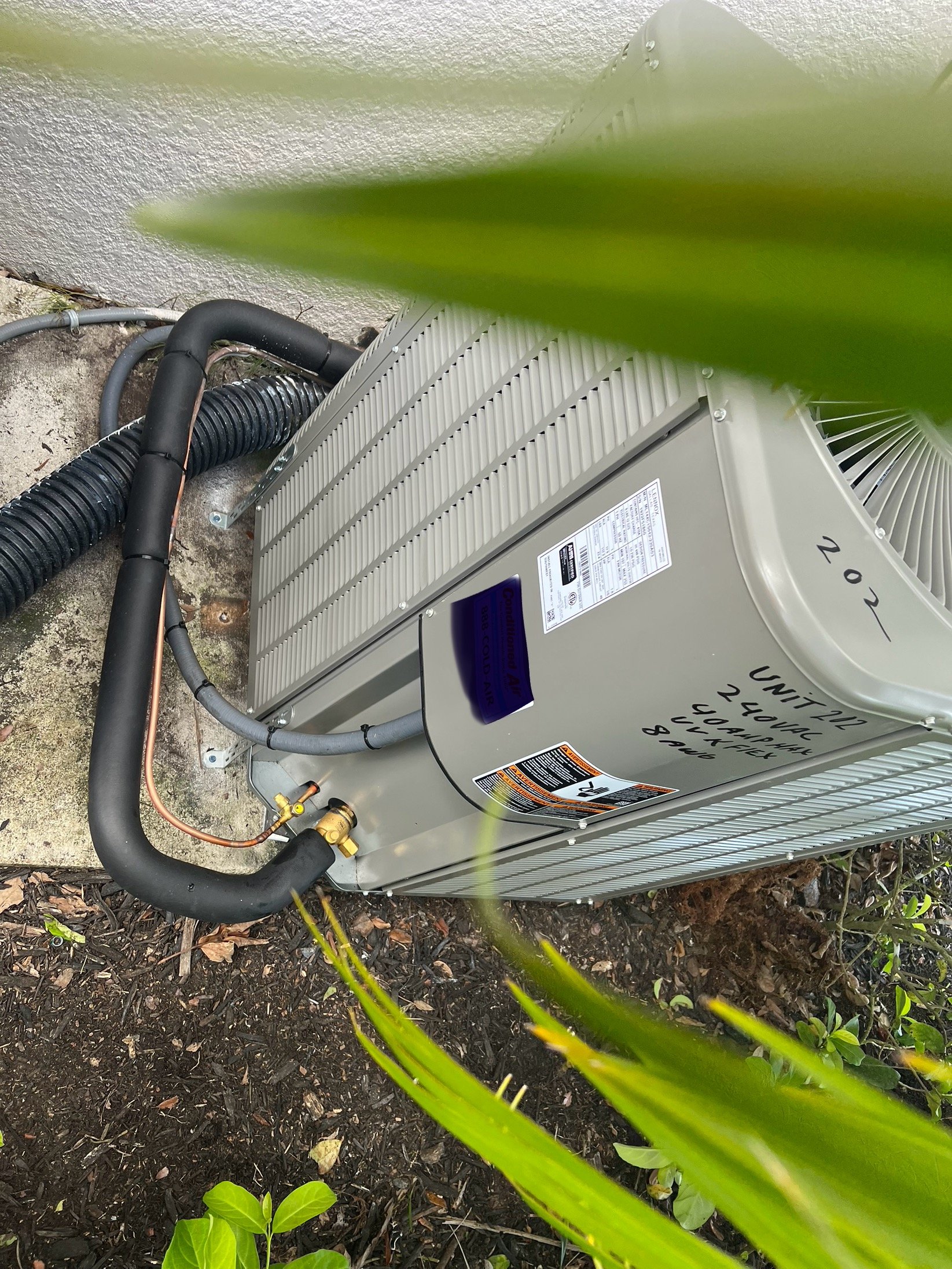 A central air conditioning unit installed outdoors, with visible pipes and wiring connected to it, situated next to a wall and surrounded by soil and plants.