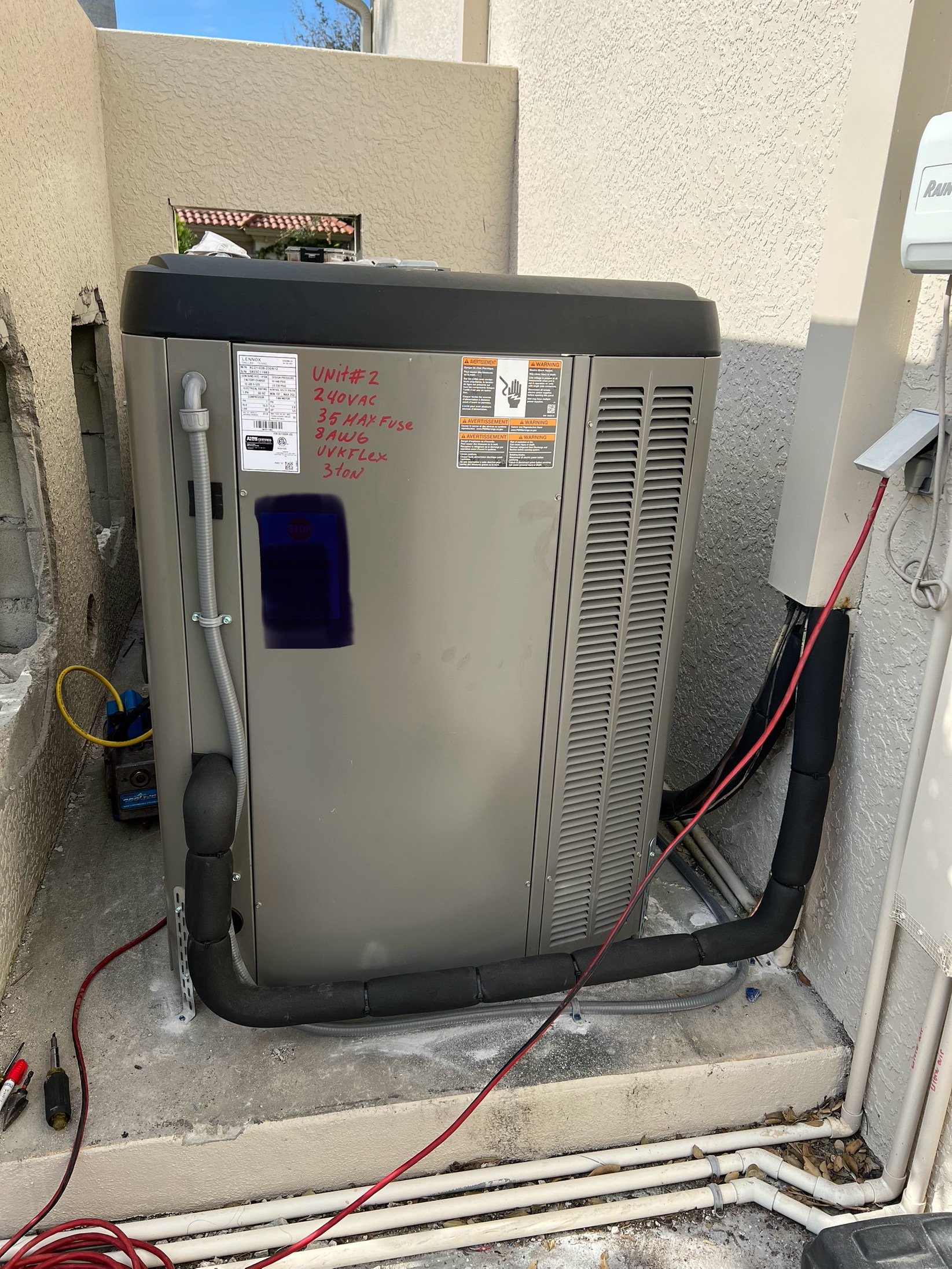 A large HVAC unit installed outside on a concrete slab on a small patio area between walls, with red and black insulated pipes and electrical wires connected to it.