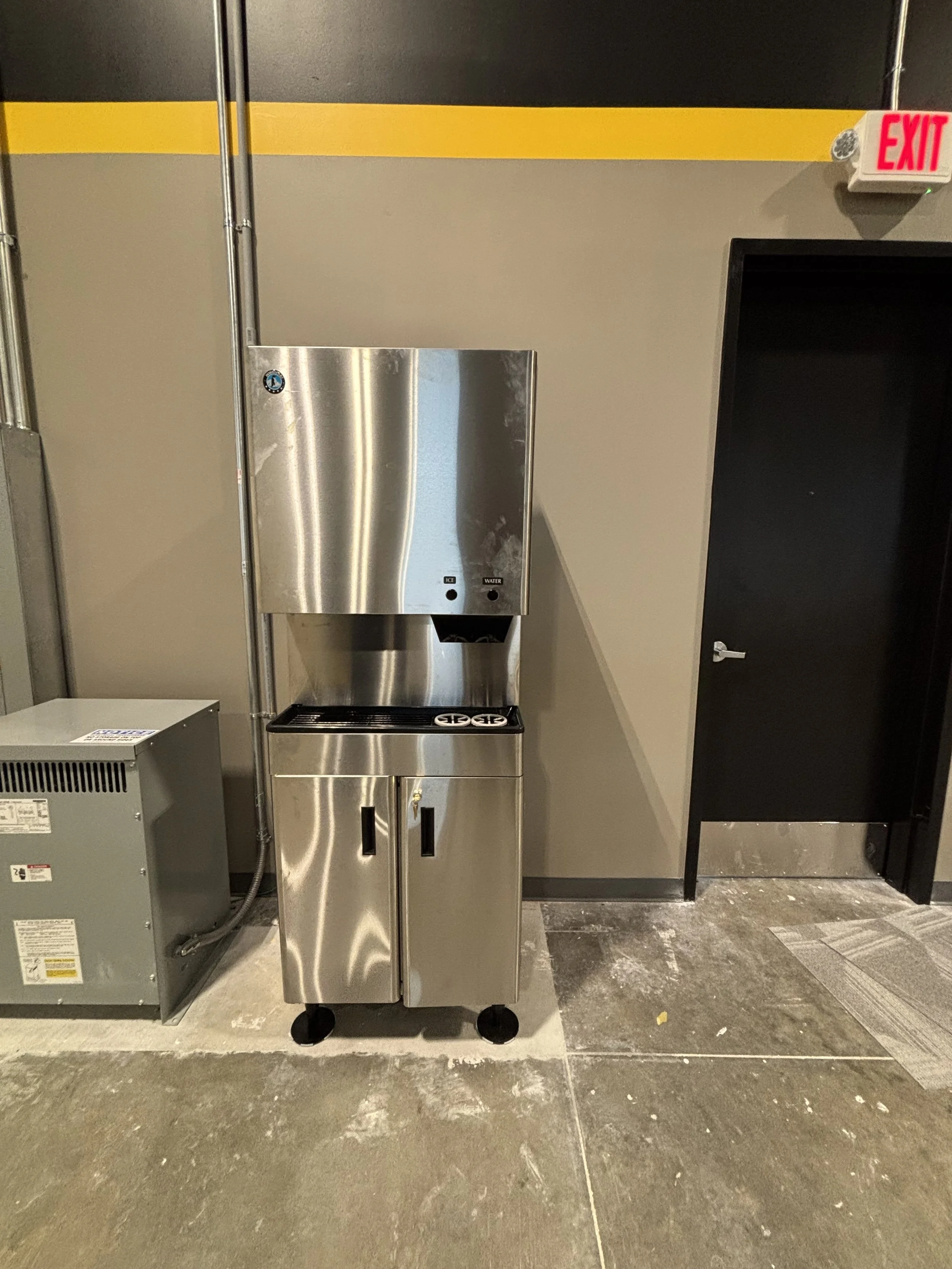Stainless steel water fountain with bottle filling station, located against a beige wall with yellow and black stripes, near a black door and an exit sign.
