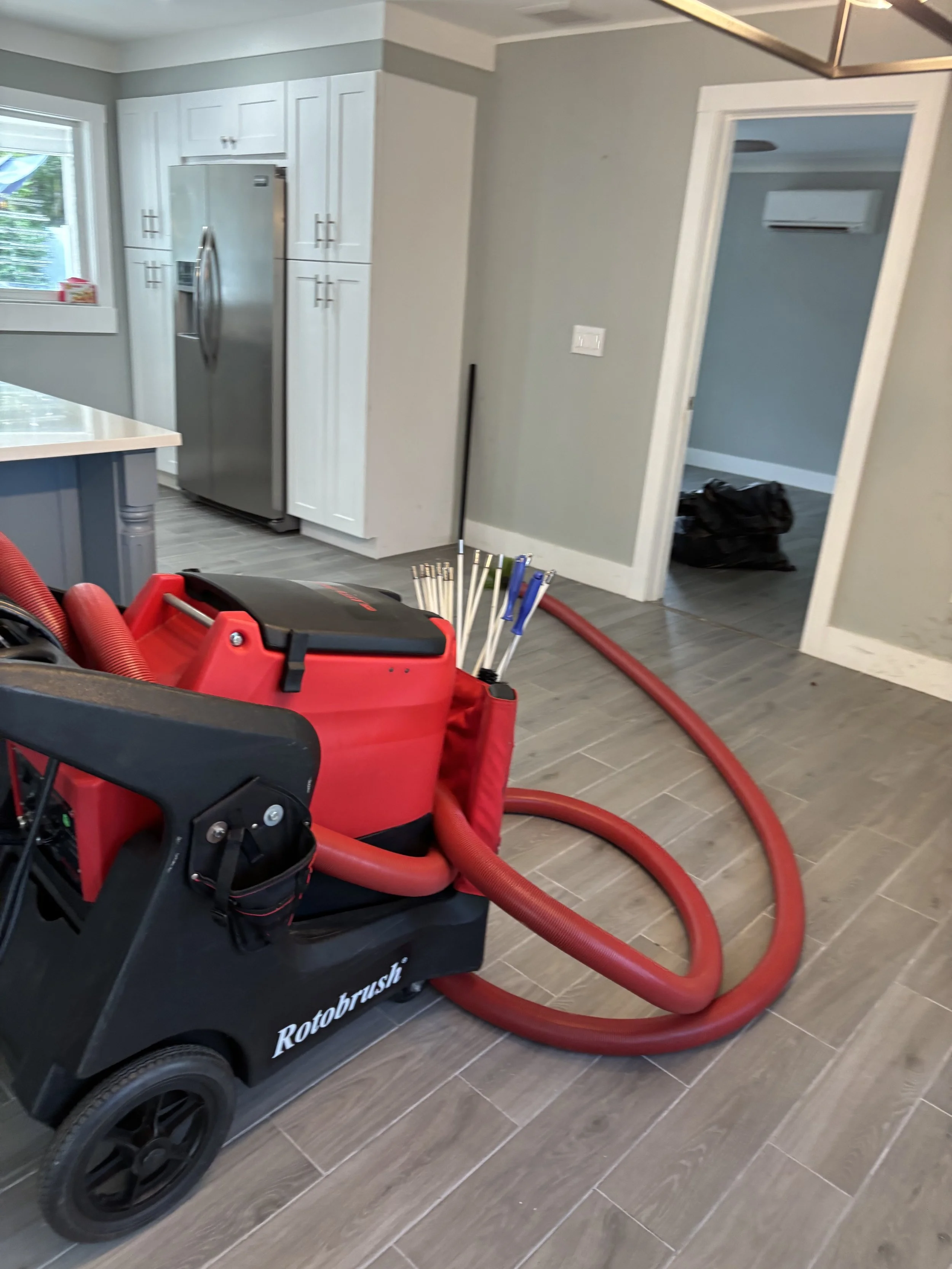 A RedRotorbrush cleaning machine with hoses and cleaning tools inside a modern kitchen with gray wood-look flooring, white cabinets, a stainless steel refrigerator, and a window with a view outside.