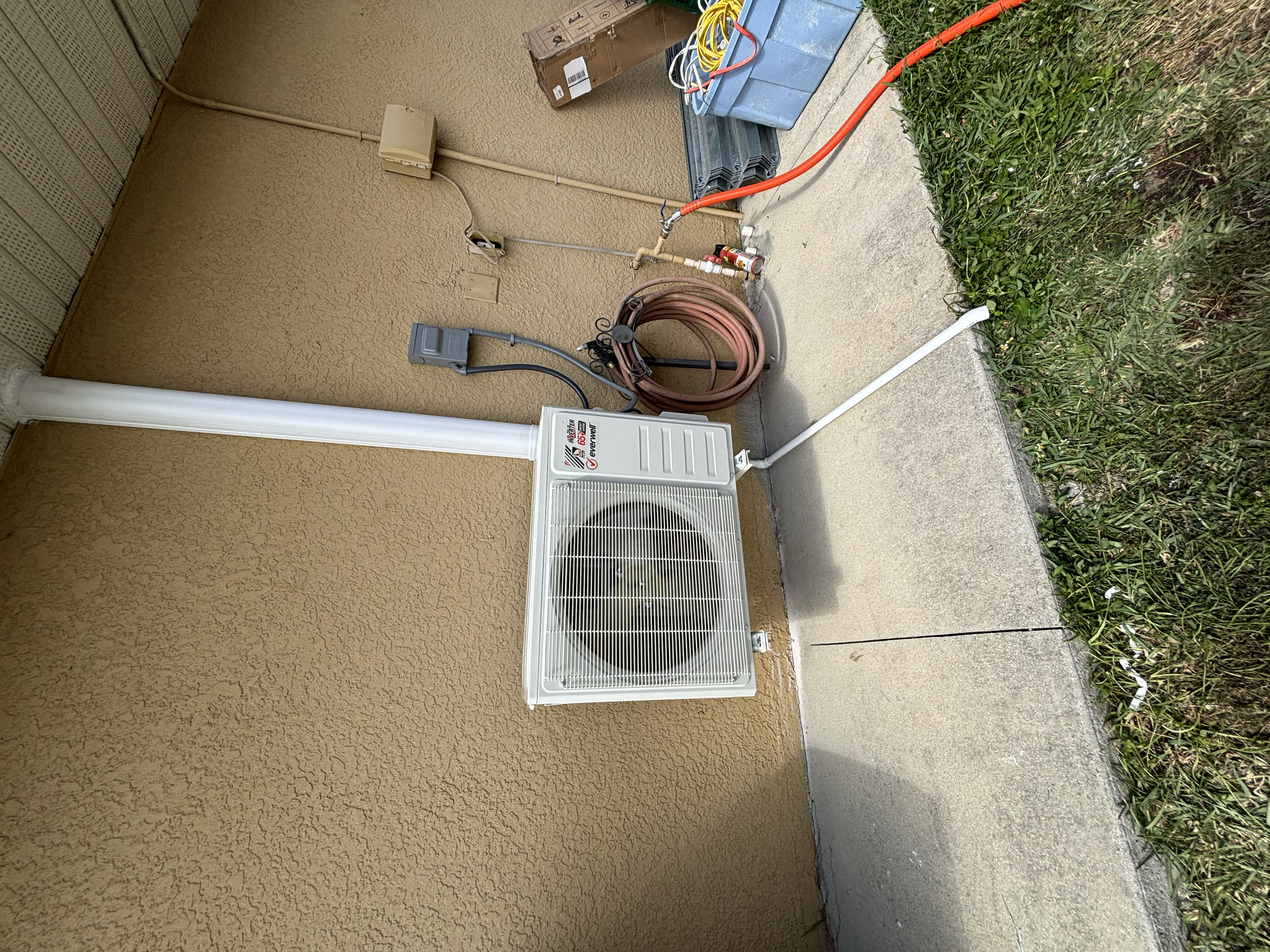 Outdoor air conditioning unit installed on a beige stucco wall, with utility boxes, electrical wiring, a garden hose coiled on the ground, and some construction or maintenance tools nearby.