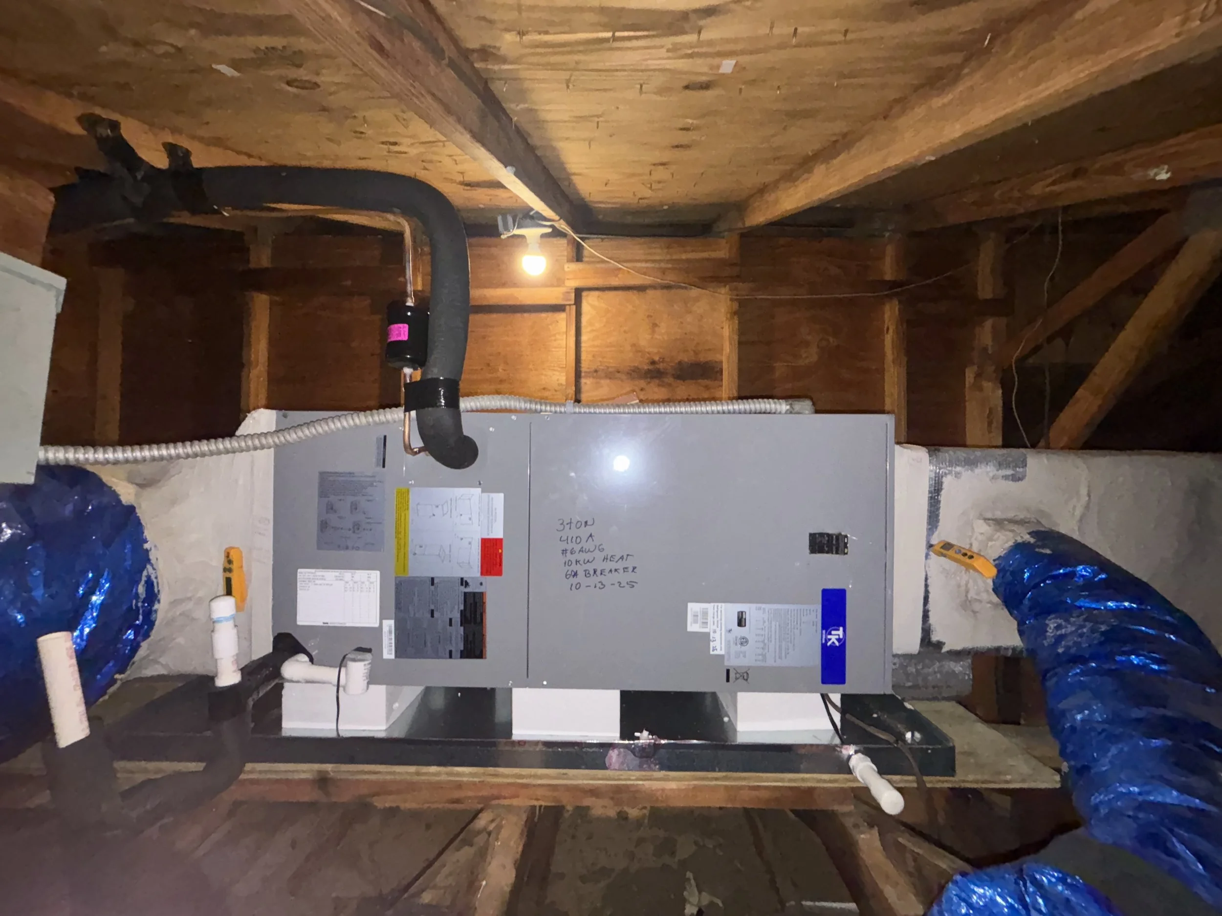 The image shows an HVAC furnace installed in an attic with wooden framing. The furnace has various ducts and cables connected to it, including a black exhaust pipe, blue insulated duct, and white PVC pipes. There is a single exposed lightbulb illumin