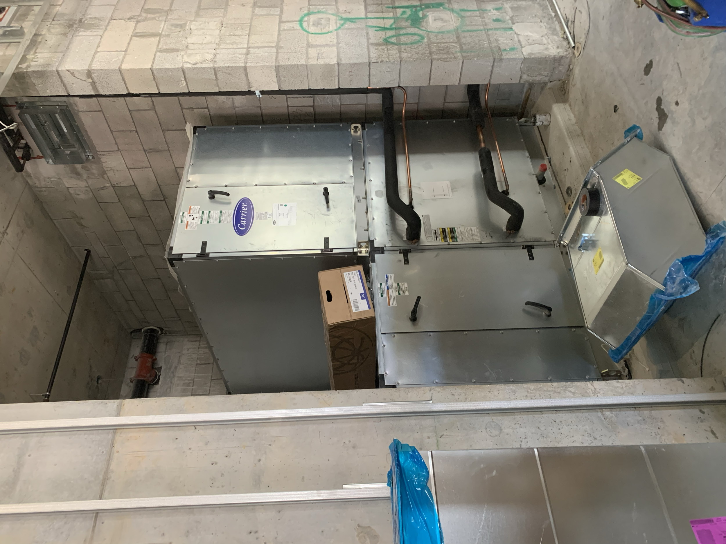 HVAC system installation in a construction site, with metal ducts, pipes, and a cardboard box on the floor.