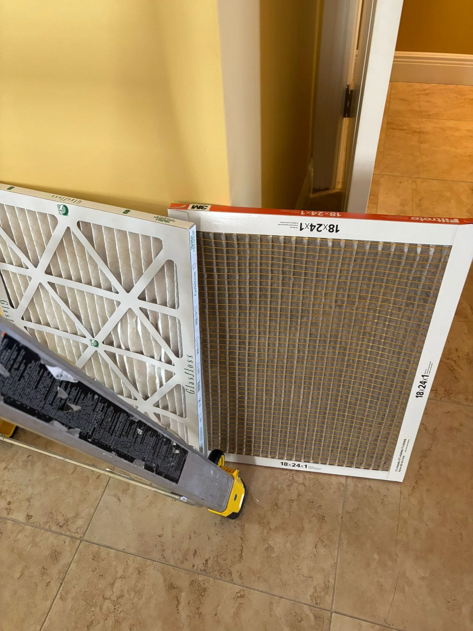 Two air filters, one with white pleated filter material and the other with a brown wire mesh, leaning against each other on a tiled floor near a yellow wall, with part of a step ladder visible in the foreground.