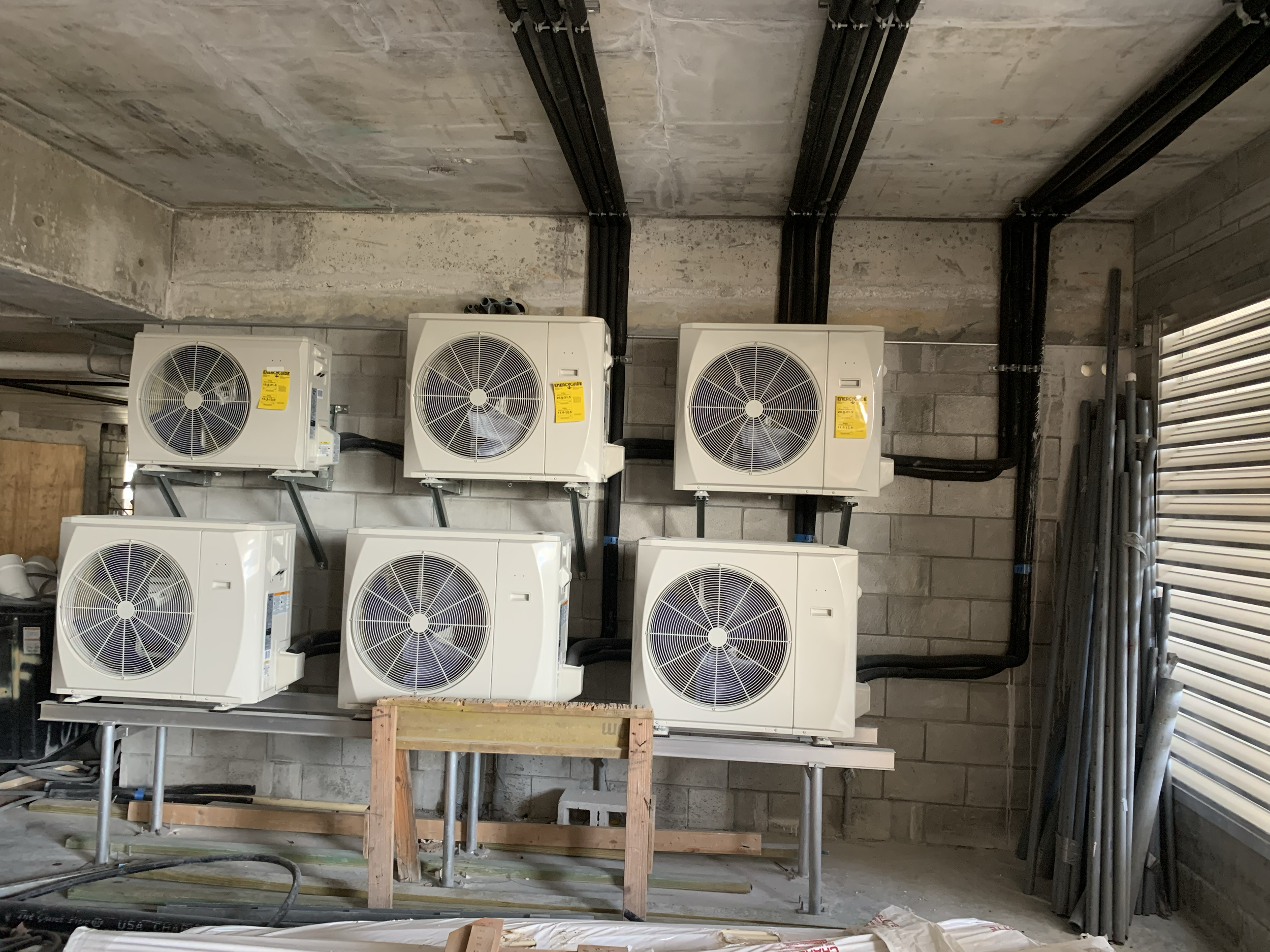 Six outdoor HVAC unit compressors mounted on a wall inside a building under construction, with black electrical conduit pipes running above and across the wall.