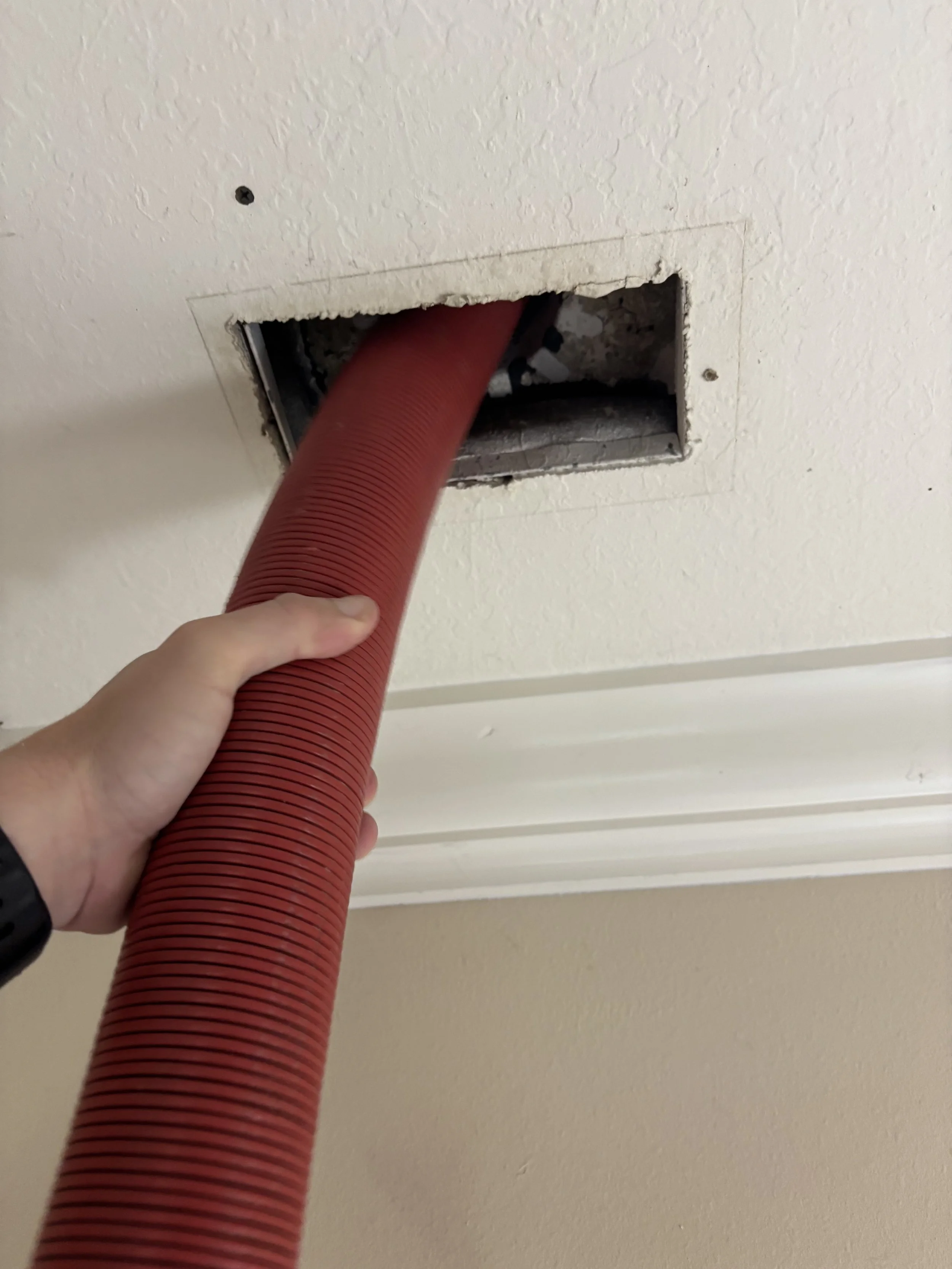 A person is holding a red flexible vacuum hose, vacuuming a ceiling vent opening in a drywall ceiling.