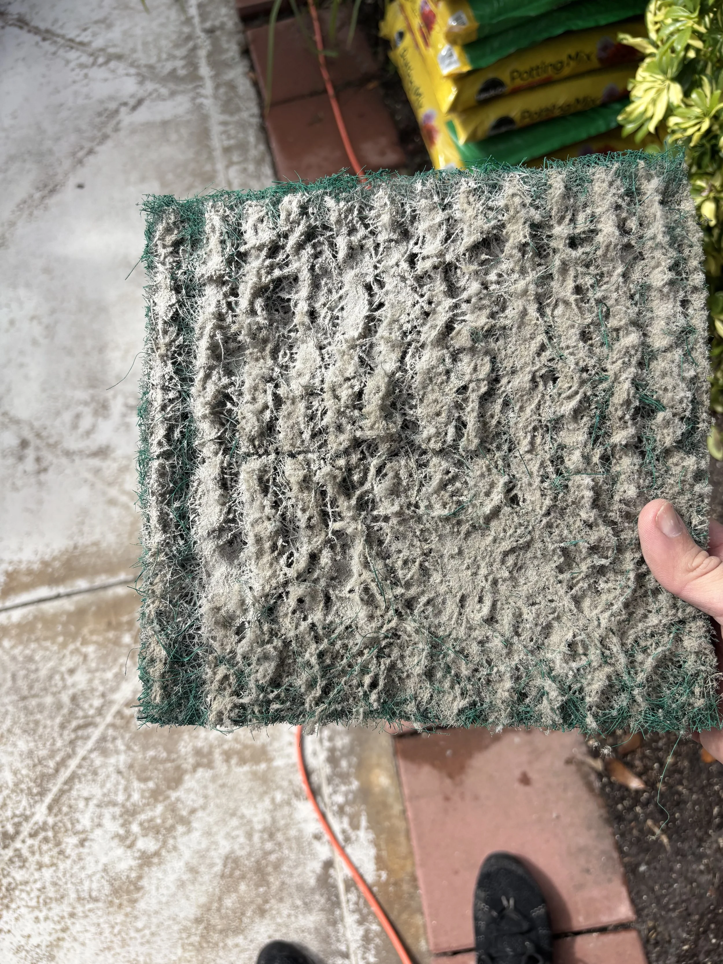 A dirty, used air filter held in a person's hand outdoors, with gardening supplies and a concrete surface visible in the background.