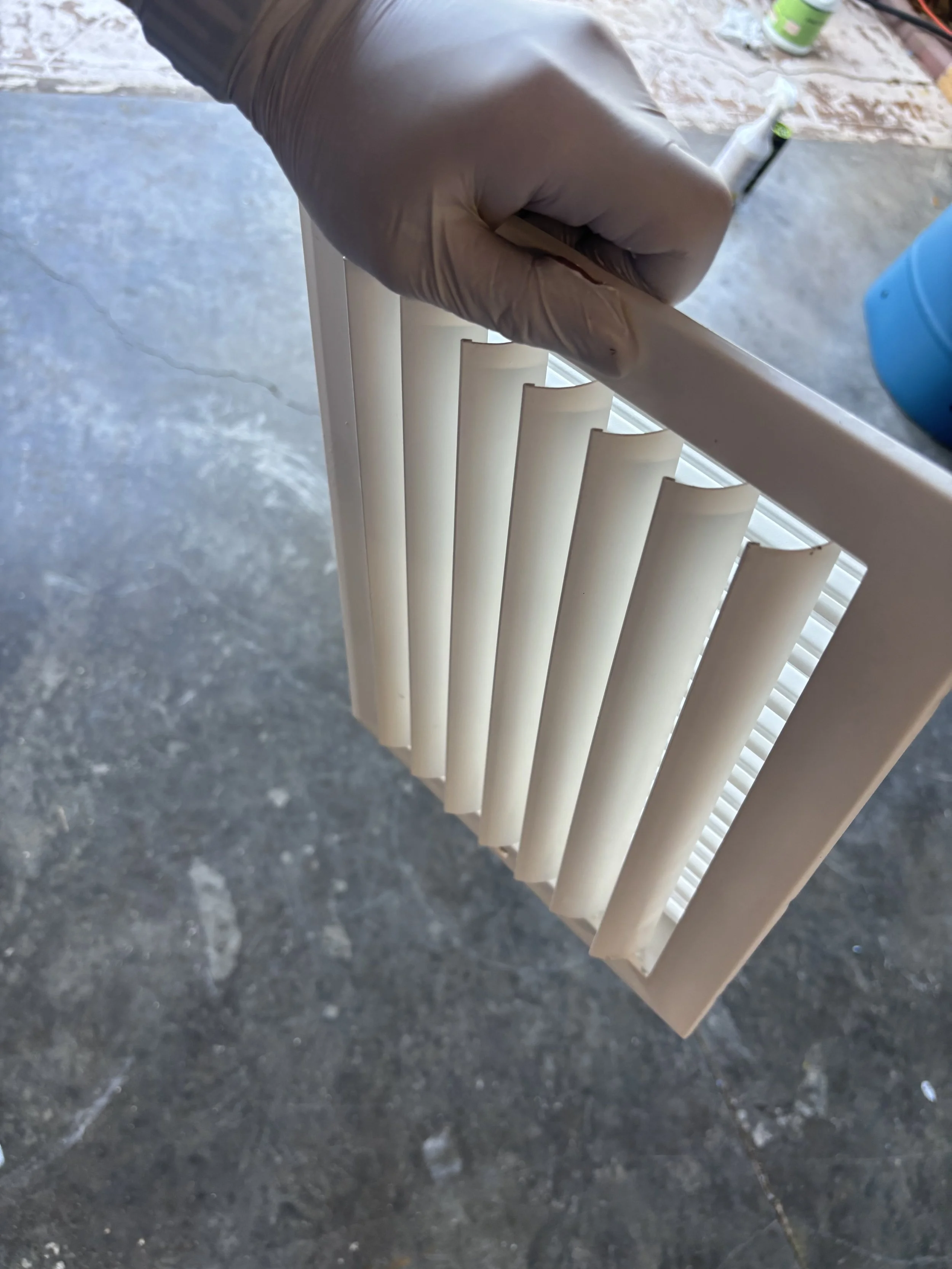 A person's hand wearing a black glove holding a white plastic window blind slat, with a gray concrete floor in the background.