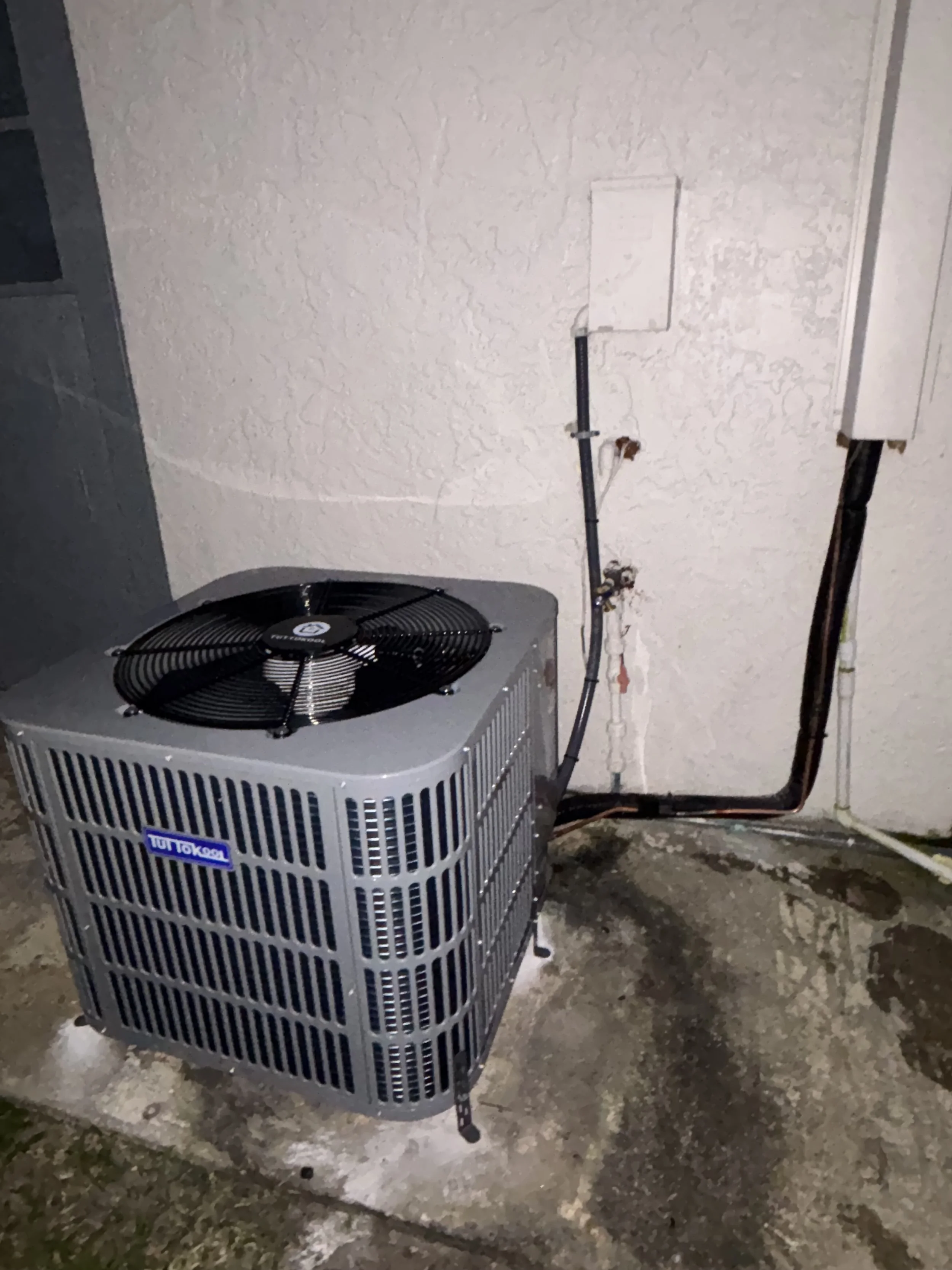 An outdoor air conditioning unit with a large fan on top, located on a concrete pad against a textured beige wall. There are black and copper pipes connected to the unit, running along the wall and into the ground.