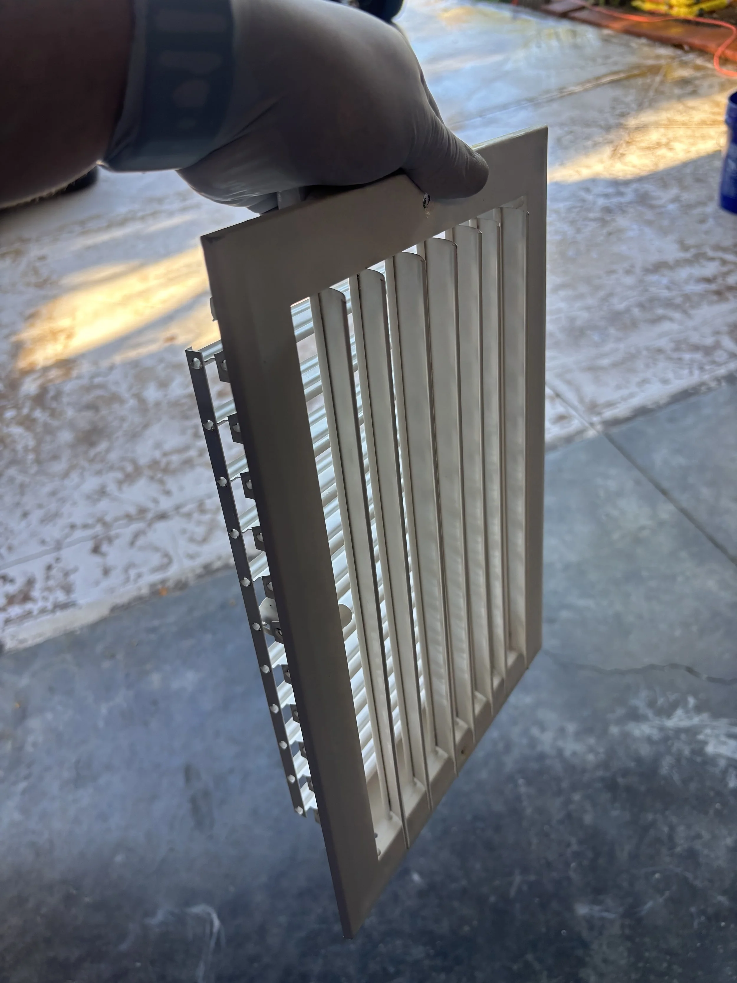 Person wearing gloves holding a metal grating or vent cover, with a concrete surface and some tools visible in the background.