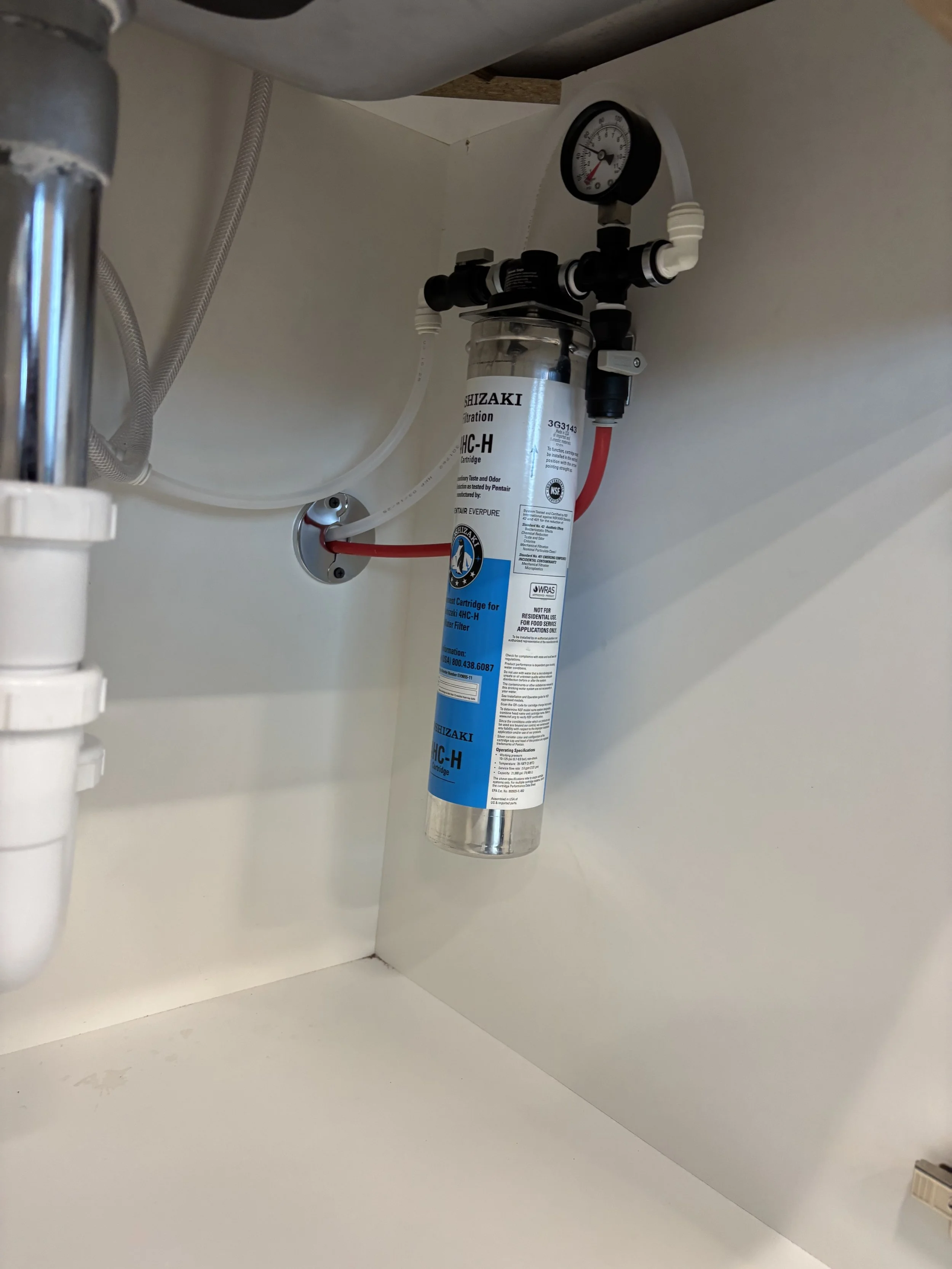 Under-sink water filtration system with a clear canister, connected to plumbing pipes, with pressure gauge on top and red and white tubes.