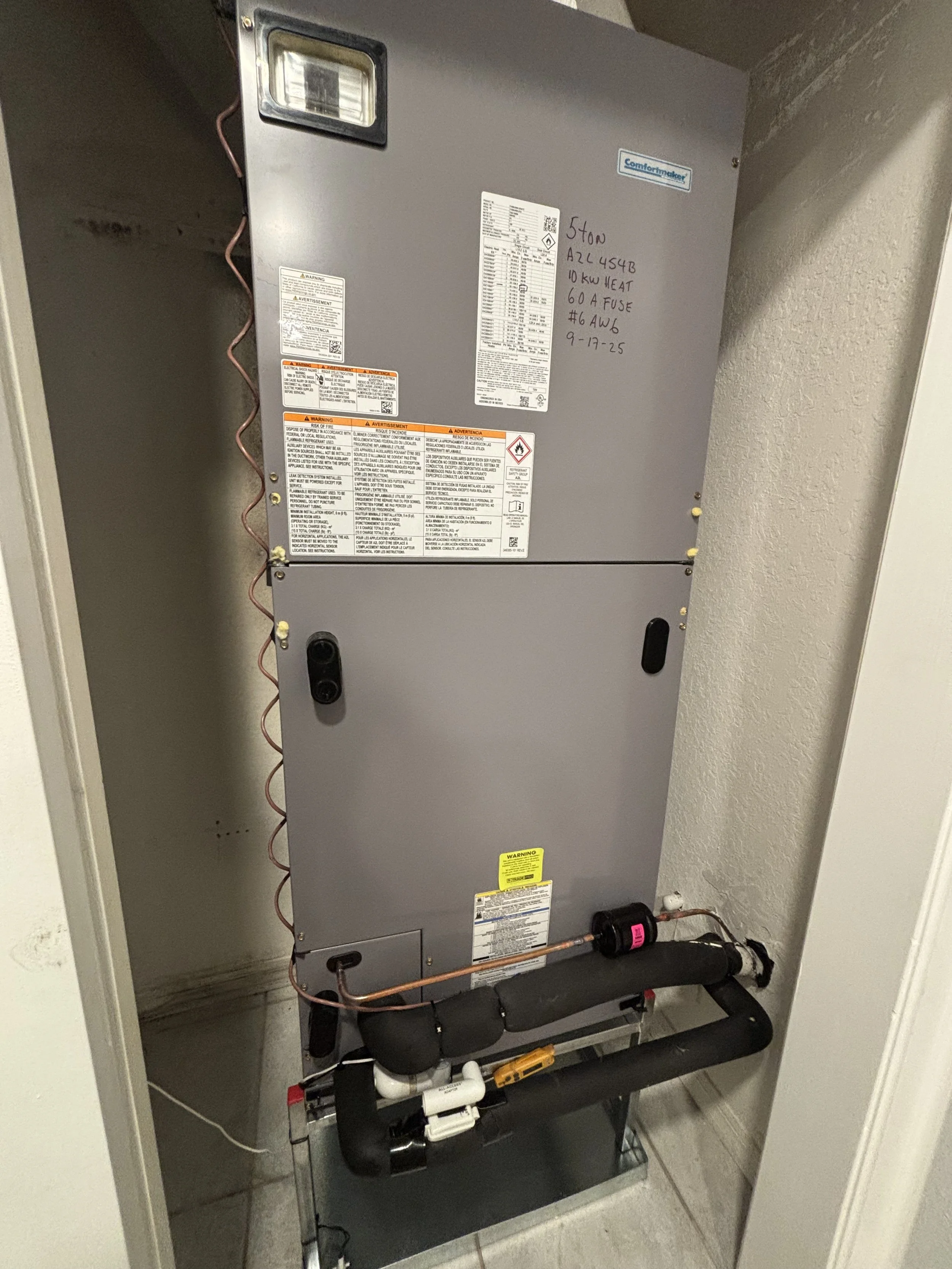 A gray HVAC or furnace unit installed in a wall niche, with warning and specification labels, some handwritten notes, and copper pipes connected at the bottom.