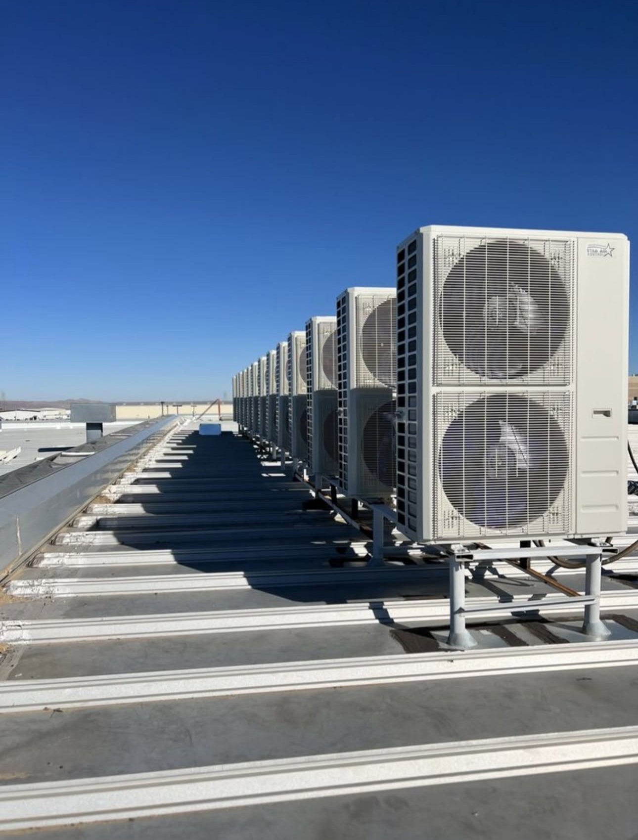 Line of multiple HVAC outdoor air conditioning units installed on a rooftop under a clear blue sky.