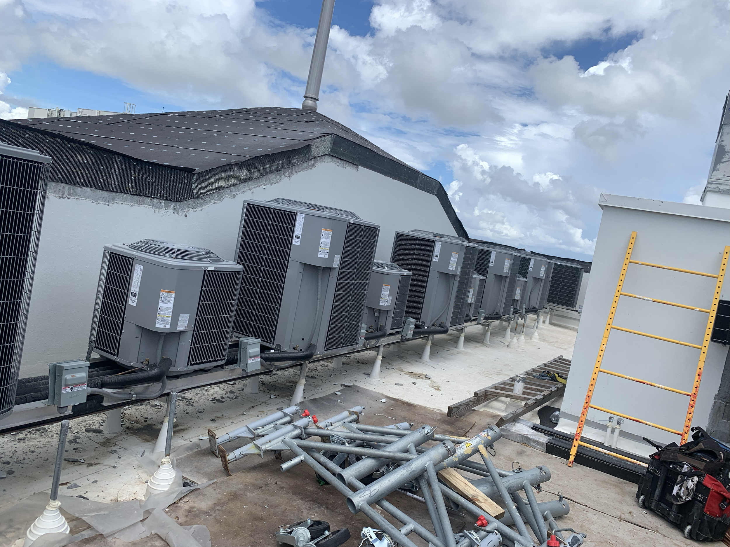 A rooftop with multiple HVAC units placed along a wall, construction tools and materials scattered, under a partly cloudy sky.