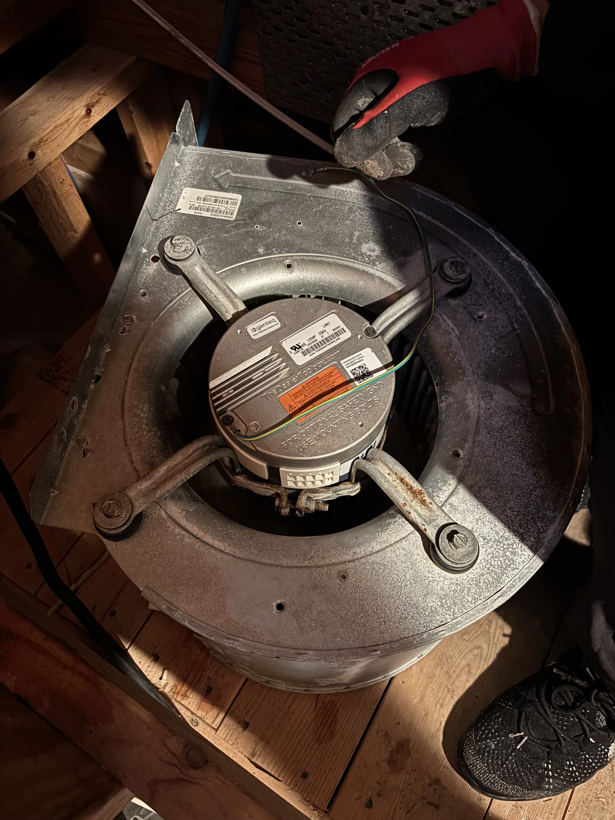 A worker's hand wearing a black and red glove holding a wire above an furnace blower wheel while installing or repairing it. The blower wheel is mounted on a metal surface, with wooden flooring underneath.