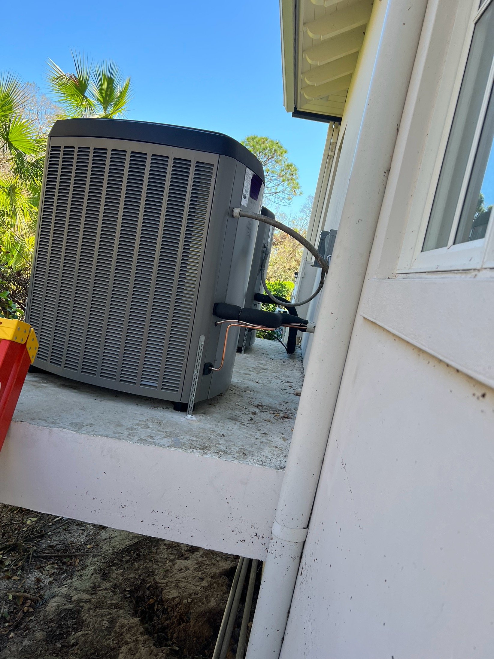 Outside view of an air conditioning unit installed on a porch next to a white house wall. Blue sky and some palm trees are in the background.