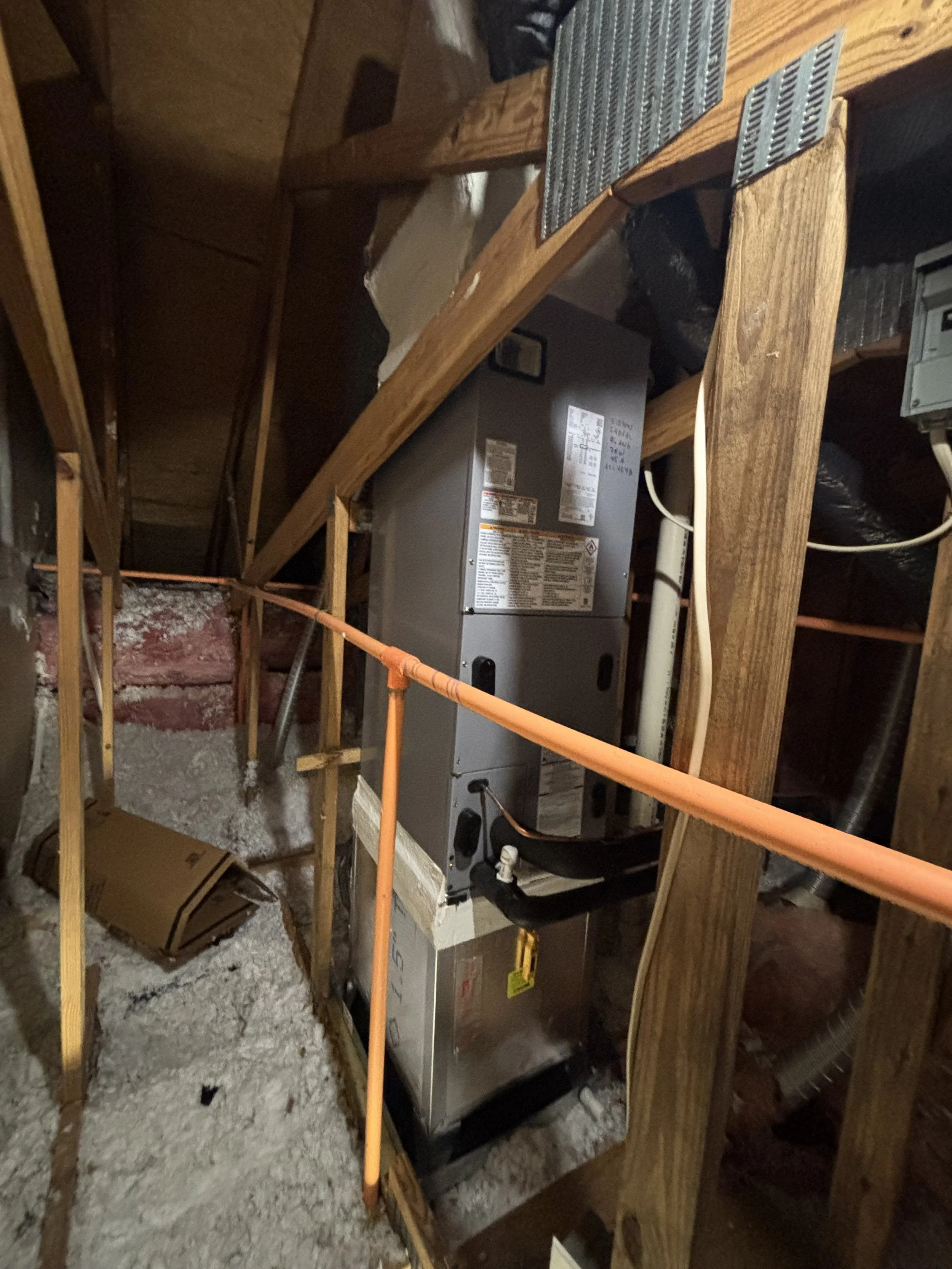 The image shows a furnace or HVAC unit installed in an unfinished attic space with insulation and wooden framing around it.