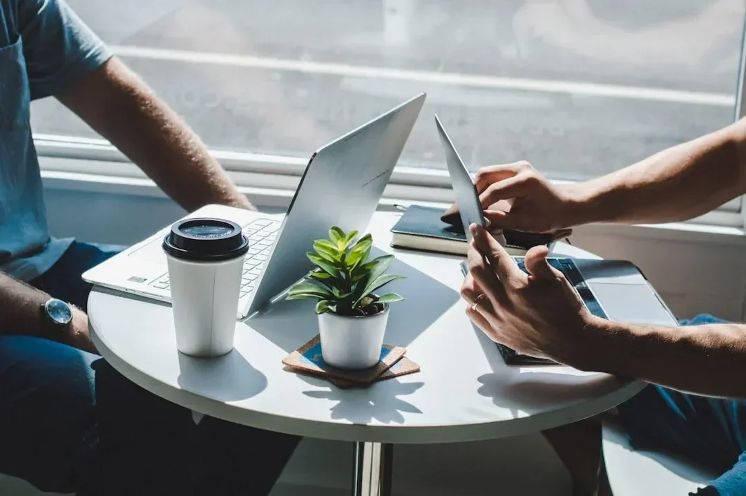 People working at a round table with laptops, tablets, notebooks, a coffee cup, and a potted plant, near large windows with sunlight.