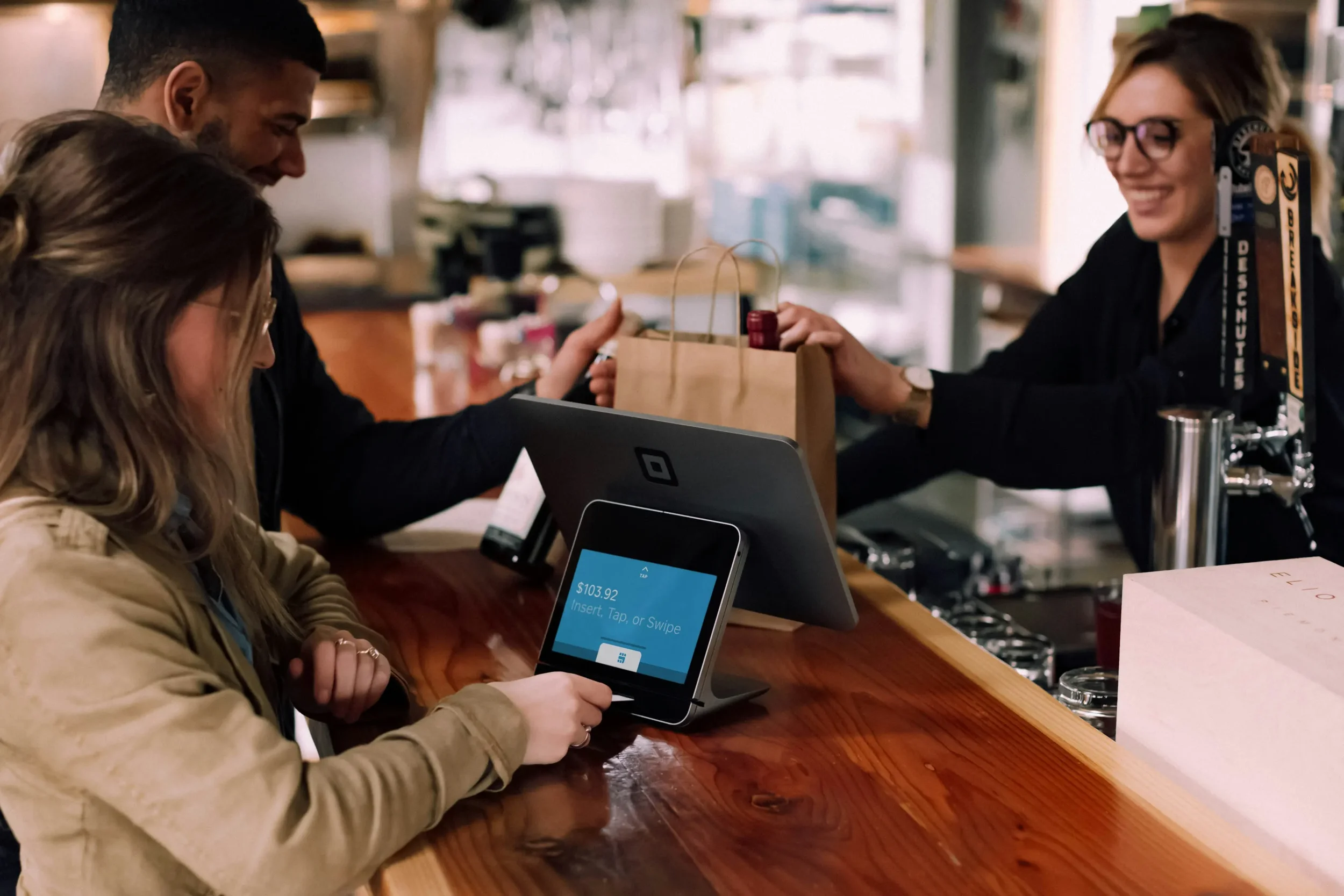 A woman sitting at a restaurant bar using a payment terminal, with a tablet displaying payment details. A man and a woman behind the bar are handing a paper bag to the woman, smiling. The woman behind the bar is wearing glasses and headphones, and the bar has a wooden countertop.