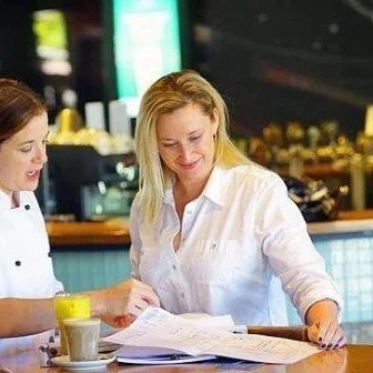 Two women sitting at a table in a café or restaurant, looking at menus and smiling.